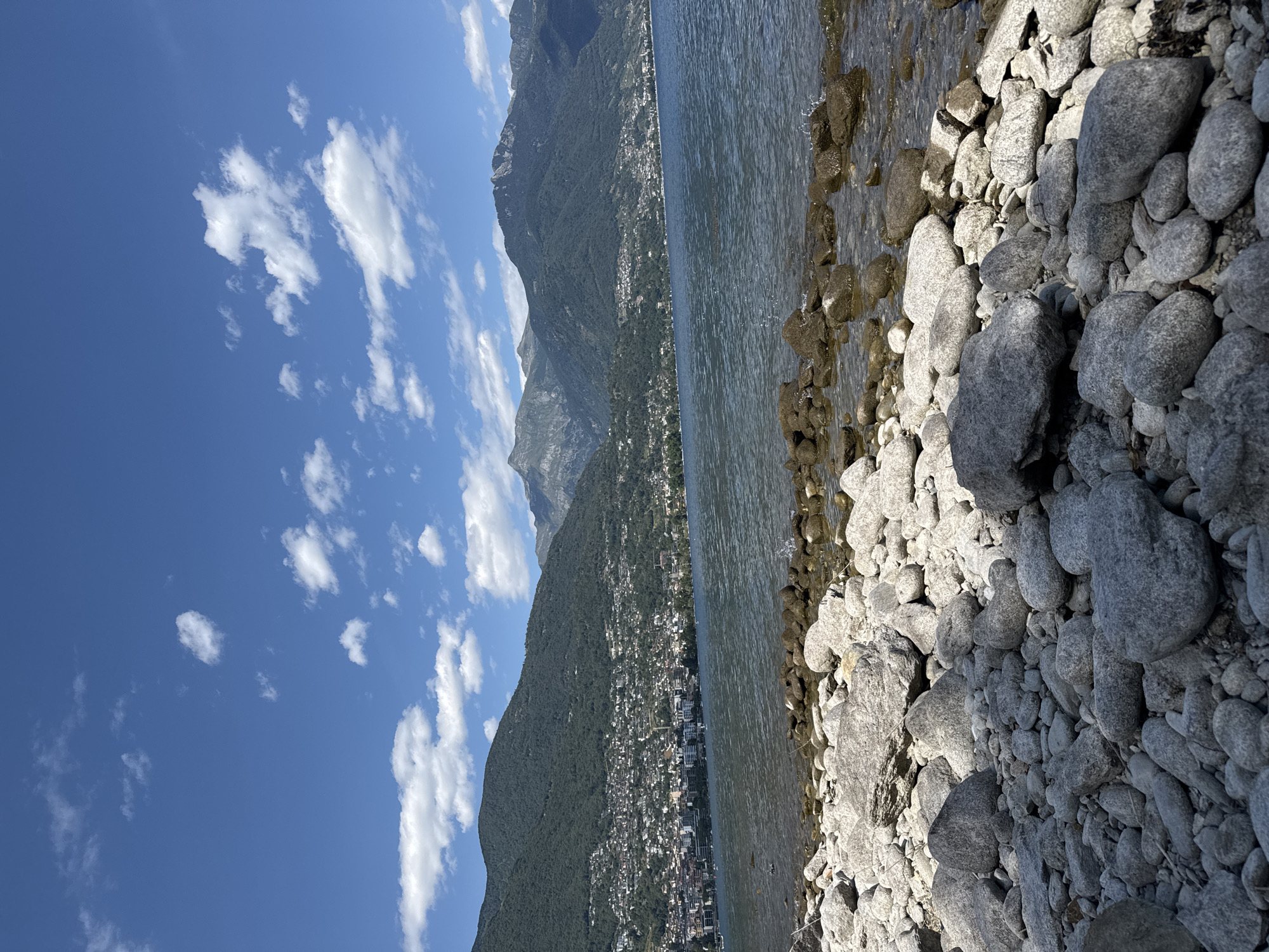 This image depicts a sunlit day at the rocky shore of a lake, likely Lake Maggiore in Locarno, Switzerland, given the context. The scene is divided into three distinct horizontal bands. The upper portion showcases a vibrant blue sky filled with scattered, fluffy white clouds, suggesting fair weather. Below the sky, a densely populated hillside slopes down towards the water. Numerous buildings with white and light-colored facades are clustered together, forming a town or city nestled amongst lush green trees. The hillsides themselves are covered in dense vegetation, indicating a warm season.
The lower part of the image is dominated by the lake's edge, characterized by a dense collection of smooth, grey and tan stones of varying sizes. The water is calm, with a gentle ripple on its surface, reflecting the sunlight and the sky. Some of the rocks are partially submerged, appearing darker and wet. The overall impression is one of tranquility and natural beauty, with the human settlement seamlessly integrated into the mountainous landscape. The lighting suggests it is daytime, likely mid-morning or afternoon, with the sun casting distinct shadows on the rocks, emphasizing their texture and form. No text is visible in the image.