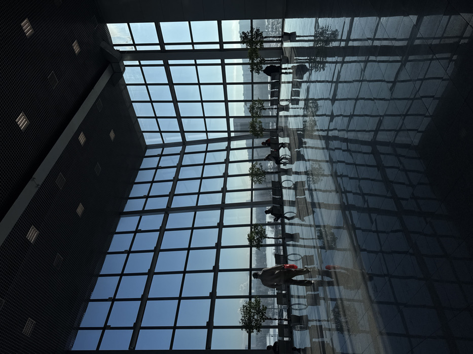 The image depicts the interior of a spacious, modern public building, likely an airport terminal, located in Khimki, Russia, consistent with Sheremetyevo International Airport. The scene is dominated by a panoramic view through two expansive, floor-to-ceiling glass curtain walls meeting at an obtuse angle, divided into a grid pattern by dark frames.
The interior features a high, dark, grid-patterned ceiling with multiple square recessed light fixtures. The floor is a highly polished, dark, reflective surface, mirroring the window grid, the silhouettes of individuals, and potted plants.
Approximately six to eight individuals are present. In the mid-ground, a male figure in light clothing walks away from the viewer towards the windows, carrying a red item in his right hand. Multiple other individuals are seated on rows of modern, bench-style seating arranged parallel to the windows. Their exact details are obscured due to strong backlighting.
A line of approximately seven potted, small trees or shrubs is positioned between the seating areas and the windows.
Through the windows, a bright, clear sky and a winter landscape are visible. This exterior view includes snow-covered ground, a body of water (possibly frozen), distant multi-story urban buildings, and a visible parking area with vehicles on the far right.
