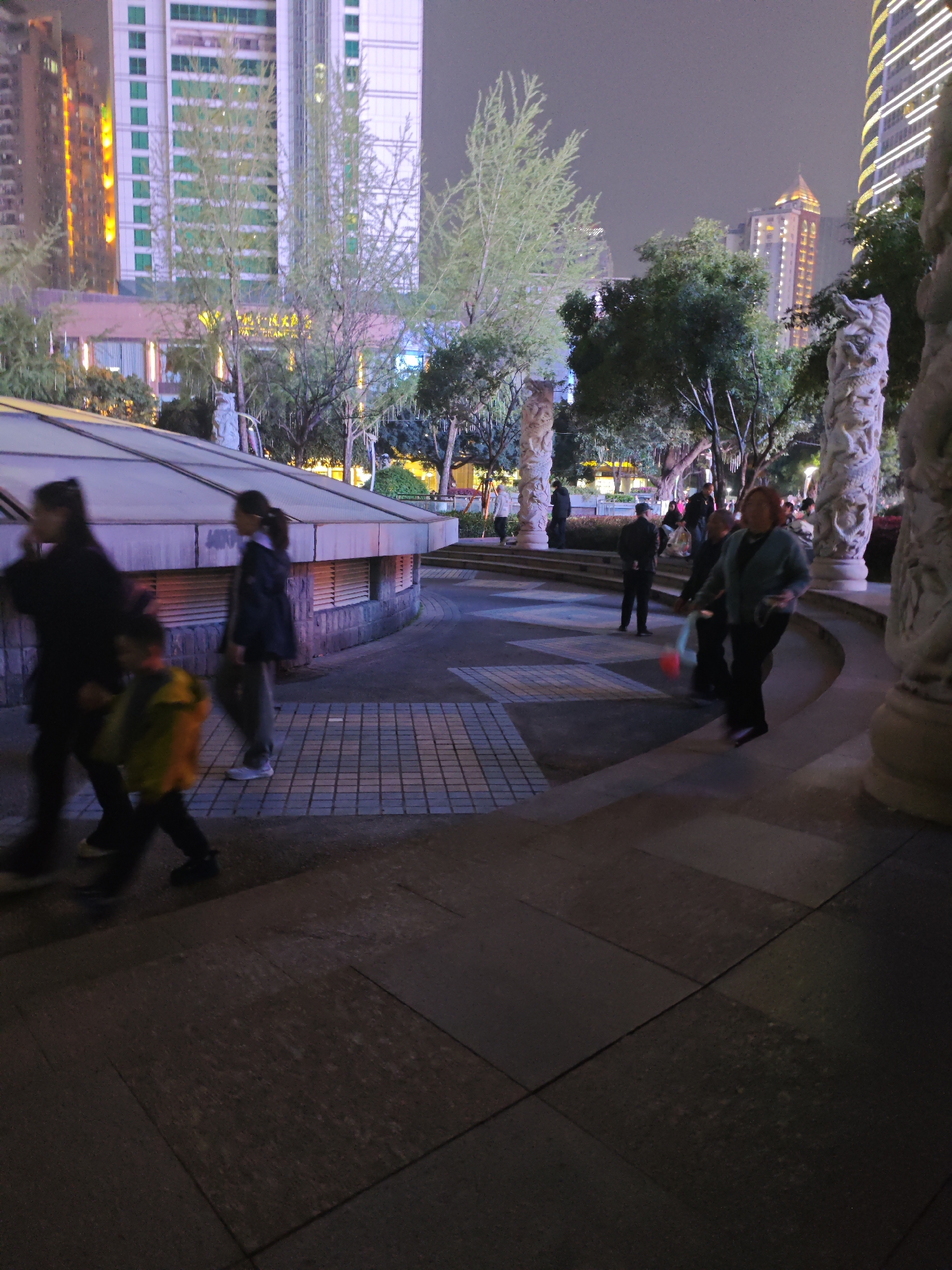 The image shows a night scene in an urban plaza in Liangjiang, China. Pedestrians, including adults and a child, are walking through the plaza. In the background, there are illuminated buildings of varying heights. Trees are interspersed throughout the scene. Elaborate stone pillars with carvings are prominent features of the plaza's architecture. The plaza has a multi-level design with steps and paved areas. Artificial lighting illuminates the area.