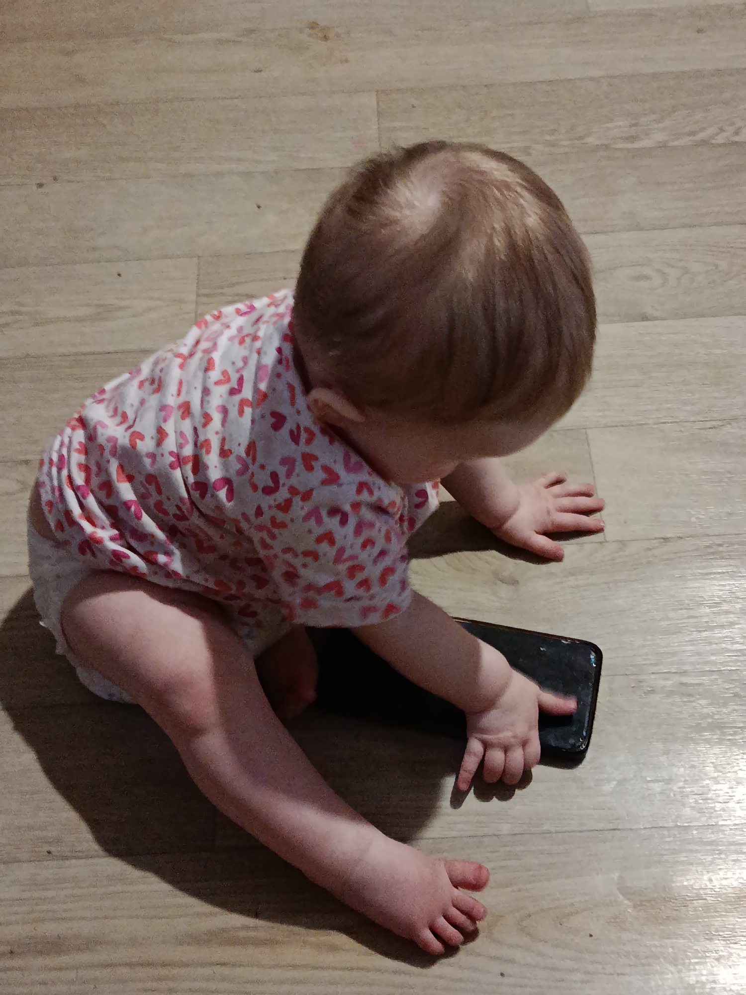 A baby is sitting on a light brown, wood-patterned floor, focusing on a black smartphone. The baby is wearing a white shirt with small pink heart patterns and a diaper. The baby is looking down at the phone, and one hand is touching the screen. The location is unknown.
