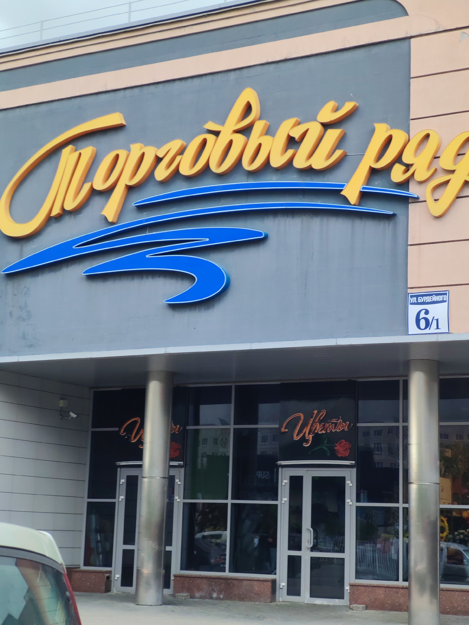 The image shows the exterior of a building with signage in Russian. Above the entrance, there is a sign that reads "Торговый ряд" in cursive yellow letters, with a blue graphic element underneath. To the right of the sign, there is a small sign indicating the address as "ул. БУРДЕЙНОГО 6/1". The entrance below features glass doors and windows, with "Цветы" (Flowers) in cursive script and a rose graphic displayed on the glass. Cylindrical silver pillars flank the entrance. A portion of a vehicle is visible on the left side of the frame. The photograph appears to be taken in an urban environment, with a cloudy sky overhead. The location is stated as Unknown, Unknown.