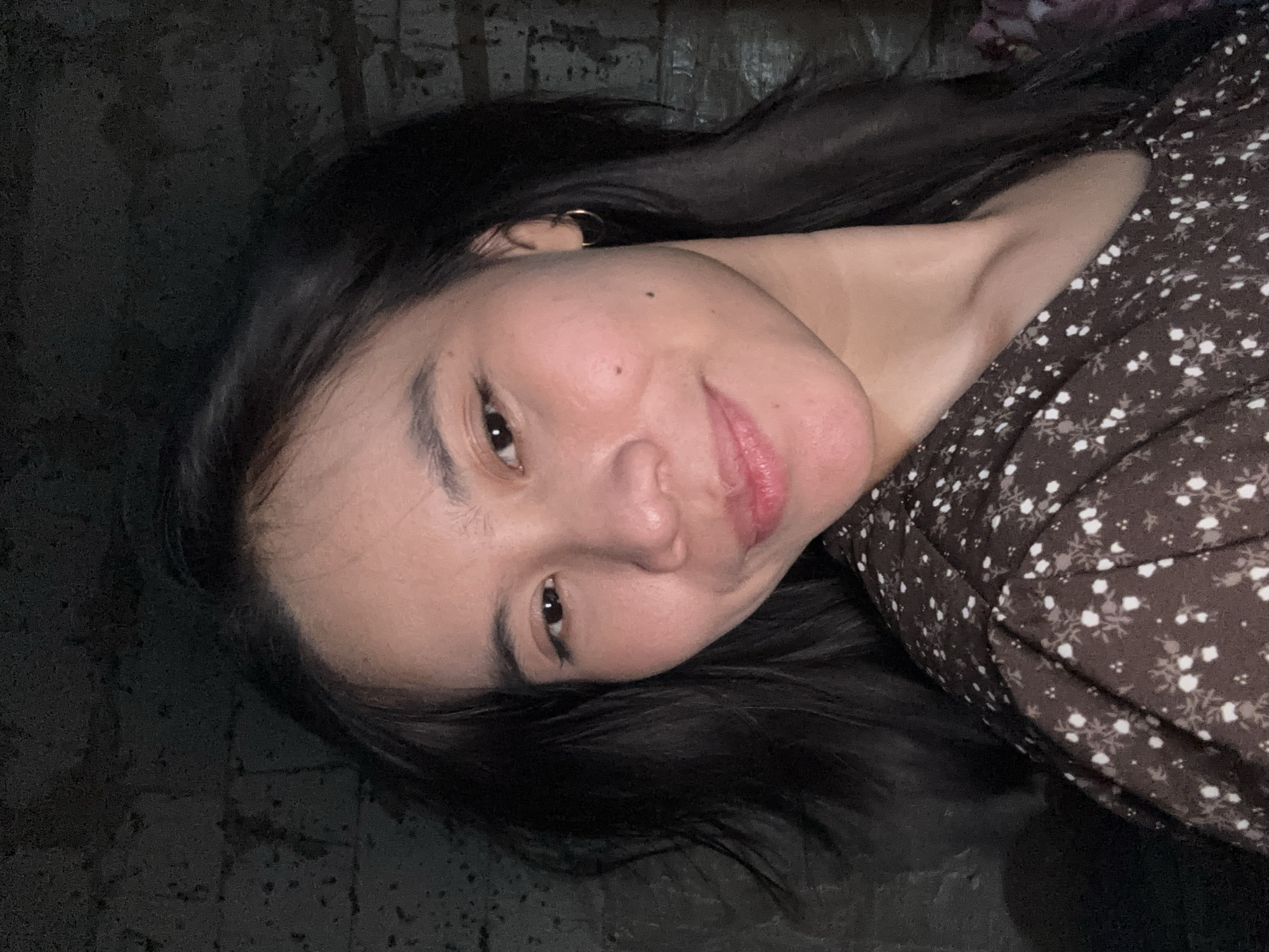 A young woman with dark hair and a brown floral dress is pictured in a close-up selfie. She is wearing a small hoop earring in her right ear. Her head is tilted slightly to her left, and she is smiling directly at the camera. The background appears to be a textured, dark brick wall. The image is not of a specific location in East Jakarta, Indonesia.