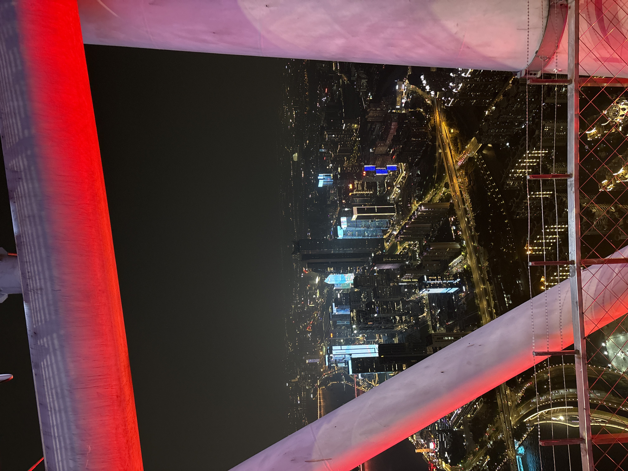 This is a nighttime aerial view of a densely populated urban area, likely a major city, focusing on the Haizhu District in China. The foreground is dominated by large, cylindrical structural elements of what appears to be an observation deck or tower, illuminated with red and pink lights. A metal railing with diamond-shaped fencing is visible at the bottom, offering a clear vantage point.
The midground and background reveal a sprawling cityscape characterized by numerous high-rise buildings, many of which are illuminated with a variety of colored lights, including blues, whites, and oranges. Highways and elevated roads crisscross the scene, with streaks of light from moving vehicles indicating traffic flow. A body of water, possibly a river or bay, is visible to the left, reflecting the city lights. The overall impression is one of a vibrant and active metropolis at night. There are no people or specific actions immediately discernible in the image. The atmosphere is dark, typical of a city at night, with the artificial lights providing the primary illumination.