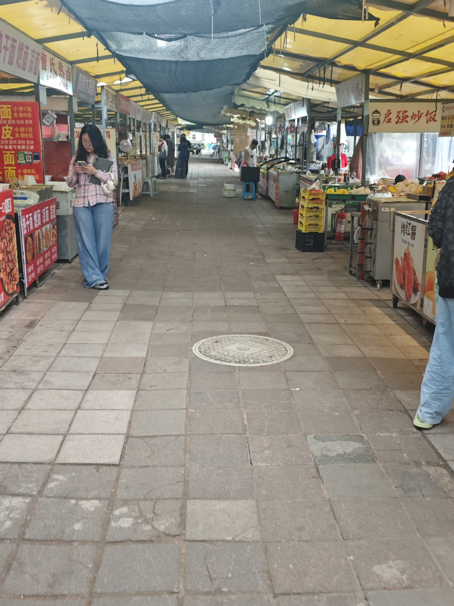 Here is a detailed description of the image:
**Location:** The image shows an outdoor market in an urban environment, likely in an area with a dense population, as indicated by the number of stalls and people. The city and specific country are unknown.
**Scene:** The main area is a wide, paved pedestrian walkway through the market. Both sides of the walkway are lined with various vendor stalls. The stalls have overhead awnings, many of which are yellow, and some are covered with dark netting. The pavement consists of gray square tiles. In the center of the walkway is a circular manhole cover.
**People:** Several people are visible in the image. A woman in a pink and blue plaid shirt and jeans stands to the left, looking at her mobile phone and holding a tablet. Other people can be seen walking further down the walkway, towards the end of the market. To the right, partially cut off by the edge of the frame, is another person in jeans. Vendors are visible at some of the stalls, and some are seated on stools.
**Objects:** The market stalls display various goods, primarily food. There are signs in Chinese characters above some stalls, and some signs display prices for food items. There are also visible crates, containers, and small carts near the stalls. On a stall to the right are food items and a red fire extinguisher.
**Actions and Interactions:** People are browsing the market stalls, and a woman is looking at her phone. Vendors are presumably selling their goods.
**Environment:** The environment suggests a typical urban marketplace with a focus on food vendors. The setting indicates a regular commercial activity.