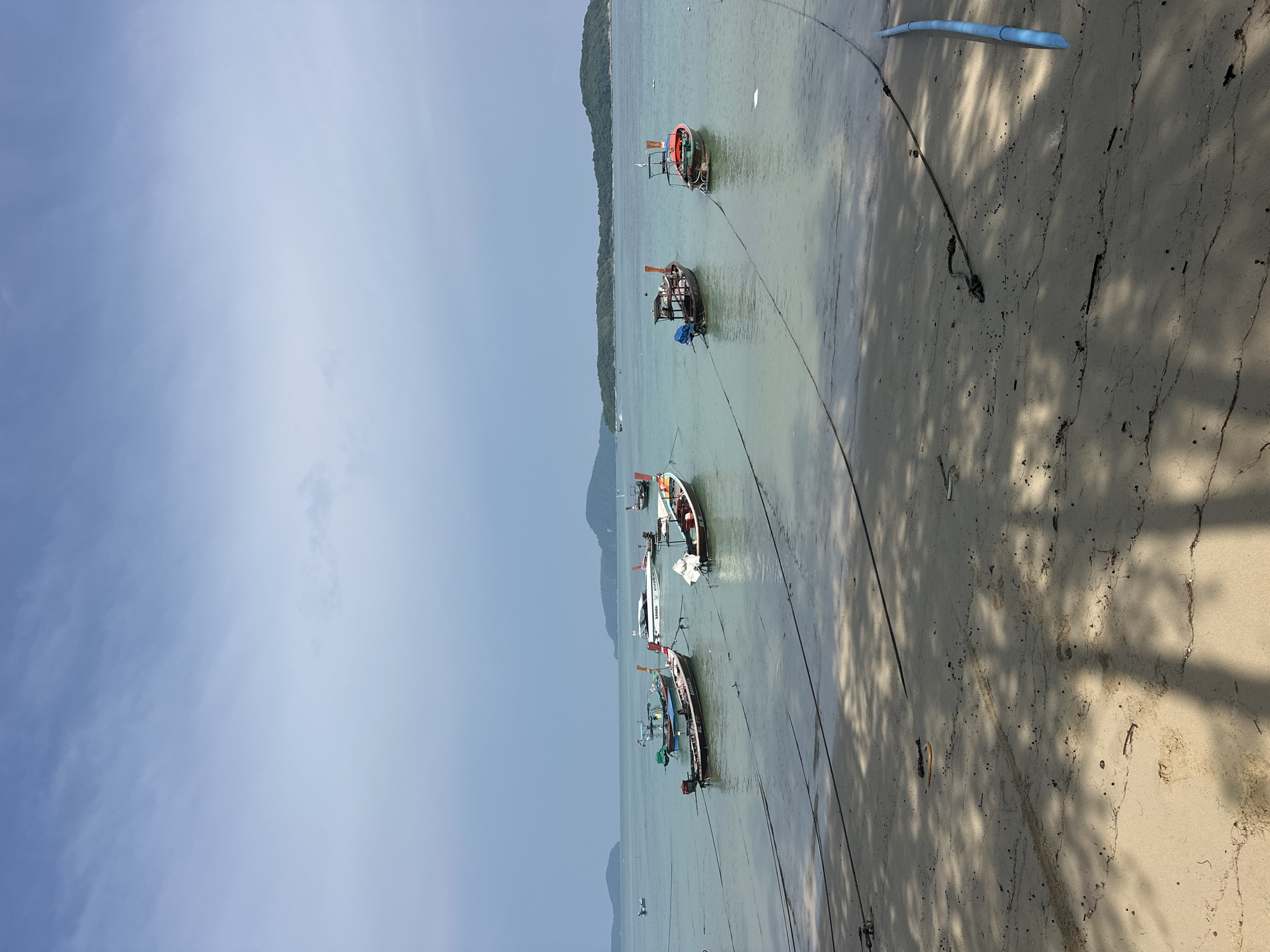 The image depicts a tranquil scene on the beach in Rawai, Thailand, with several longtail boats moored in the shallow, clear turquoise water. The sand in the foreground is a light tan color, marked with tire tracks and scattered debris, and dappled with the shadows of overhead trees. In the distance, lush green islands rise from the horizon, forming a picturesque backdrop to the seascape. The sky is a pale blue with wispy clouds, suggesting a calm, possibly overcast day. The boats, characteristic of Thailand's coastal regions, are of various sizes and colors, some with wooden hulls and distinctive long poles extending from their sterns. Their mooring ropes create a network of dark lines stretching from the shore into the water, anchoring them in place. The overall atmosphere is one of peaceful serenity, with the gentle lapping of waves and the stillness of the moored vessels conveying a sense of calm and quiet.