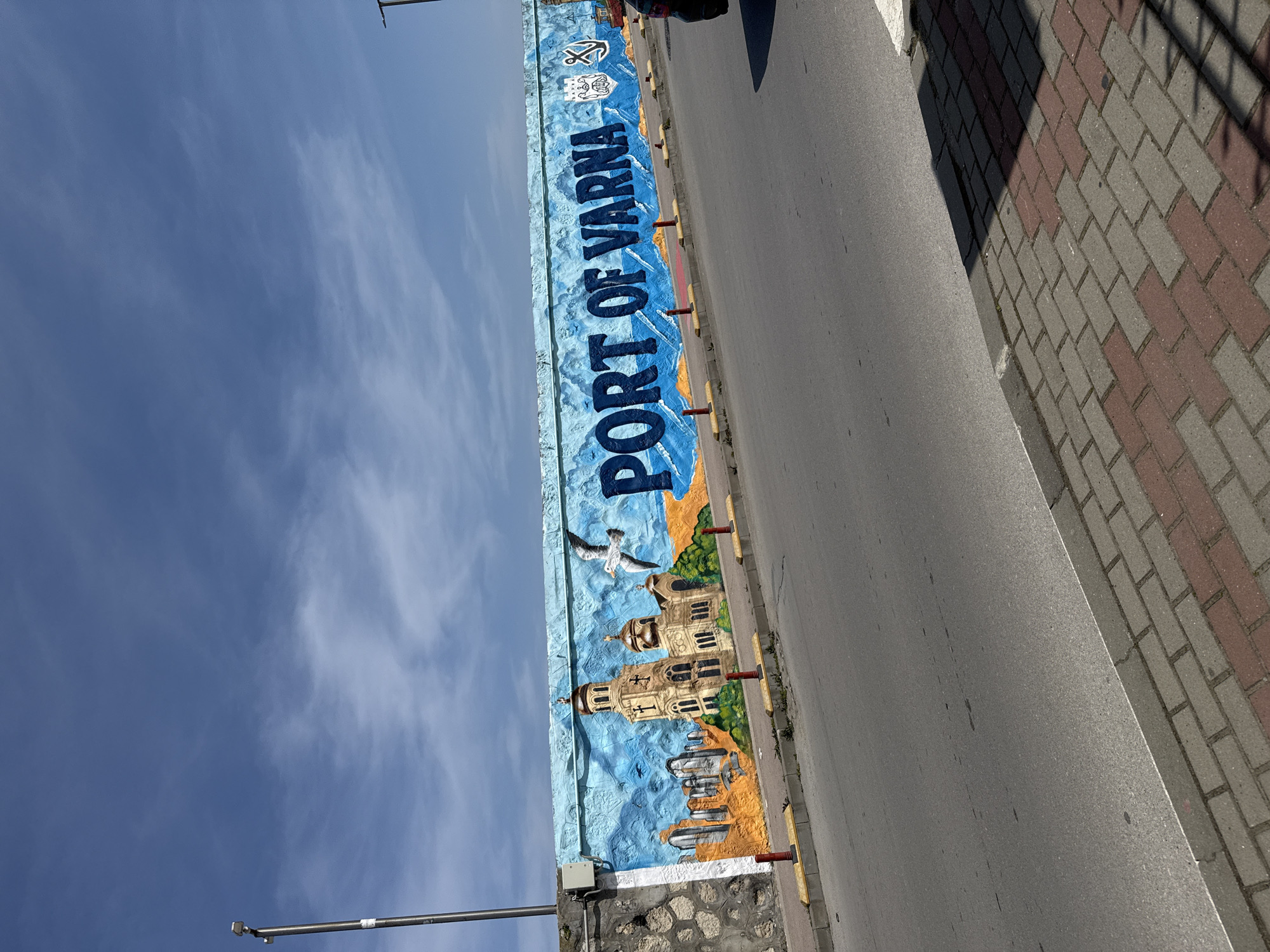 The image shows a mural along a street in Varna, Bulgaria. The mural is painted on a retaining wall or large concrete barrier. It features the words "PORT OF VARNA" in blue letters, along with an illustration of the Varna Cathedral, seagulls, and maritime symbols. The sky is visible above the mural, with a few clouds. The street and a sidewalk made of patterned bricks are adjacent to the mural. A light post is visible.