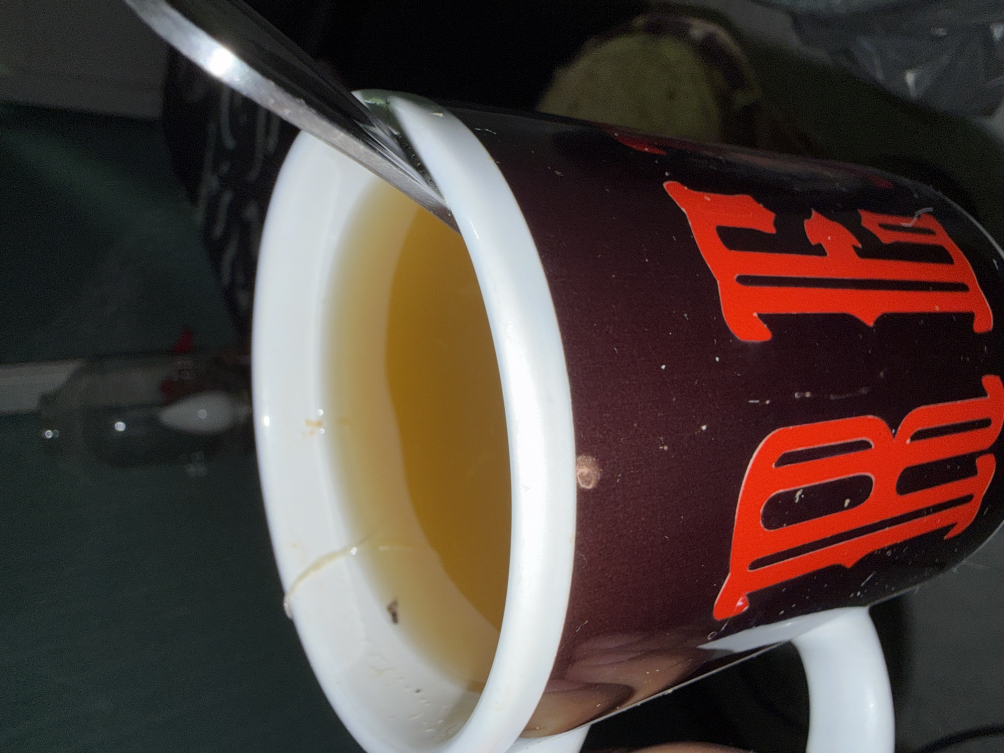 A close-up, angled shot depicts a maroon mug with red lettering. A silver spoon rests partially inside the mug, which is filled with a yellow liquid. The mug has a white handle and rim. The background is blurred, suggesting a domestic environment. There is no indication of the location in Domont, France in the image.