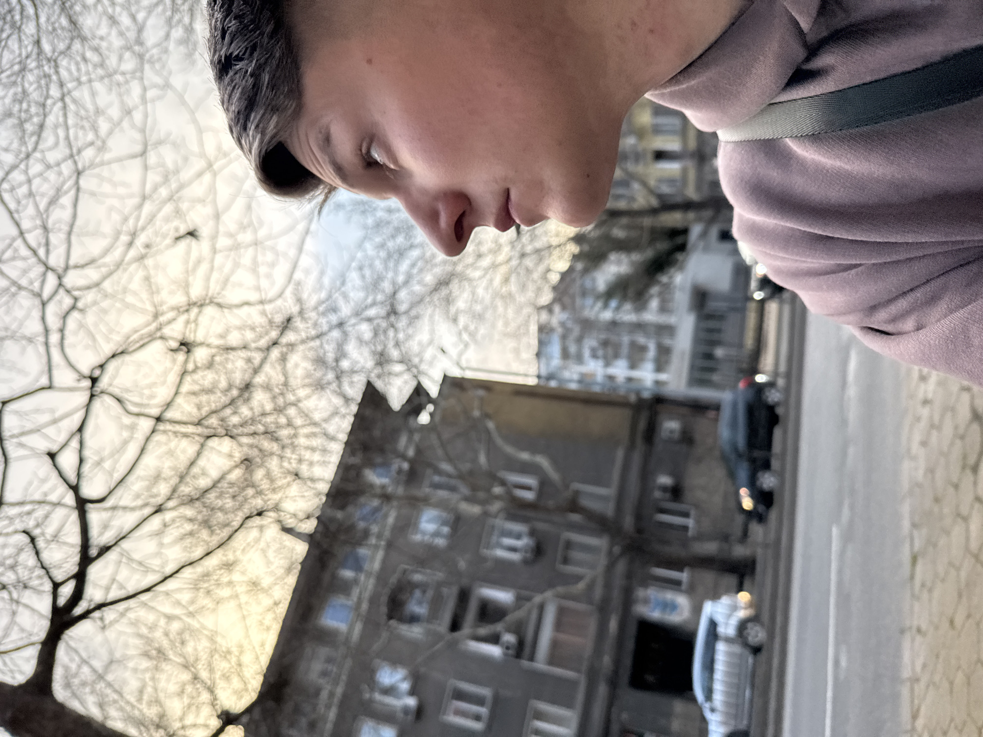 Close-up, skewed angle shot of a person's head and shoulders, seen in profile against an urban background. The person has short, dark hair and is wearing a light-colored hoodie with a strap visible on the shoulder. The background includes bare tree branches, a multi-story building with visible windows, parked cars, and a sidewalk. The scene suggests an urban environment, possibly in Plovdiv, Bulgaria, given the style of architecture and street layout.