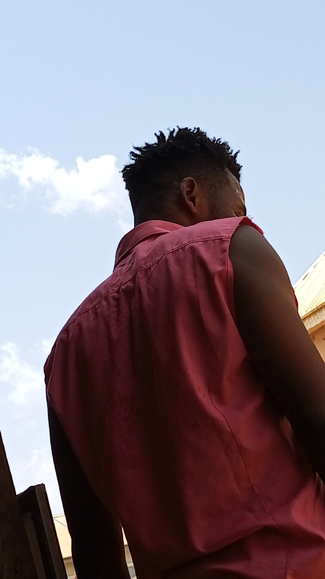 A low-angle shot captures the back of a dark-skinned person wearing a red sleeveless shirt. Their short, dark hair is visible. The background reveals a blue sky with scattered white clouds. In the lower portion of the frame, portions of buildings can be seen. It is not possible to confirm that this is Sabon Tasha, Nigeria without more context.