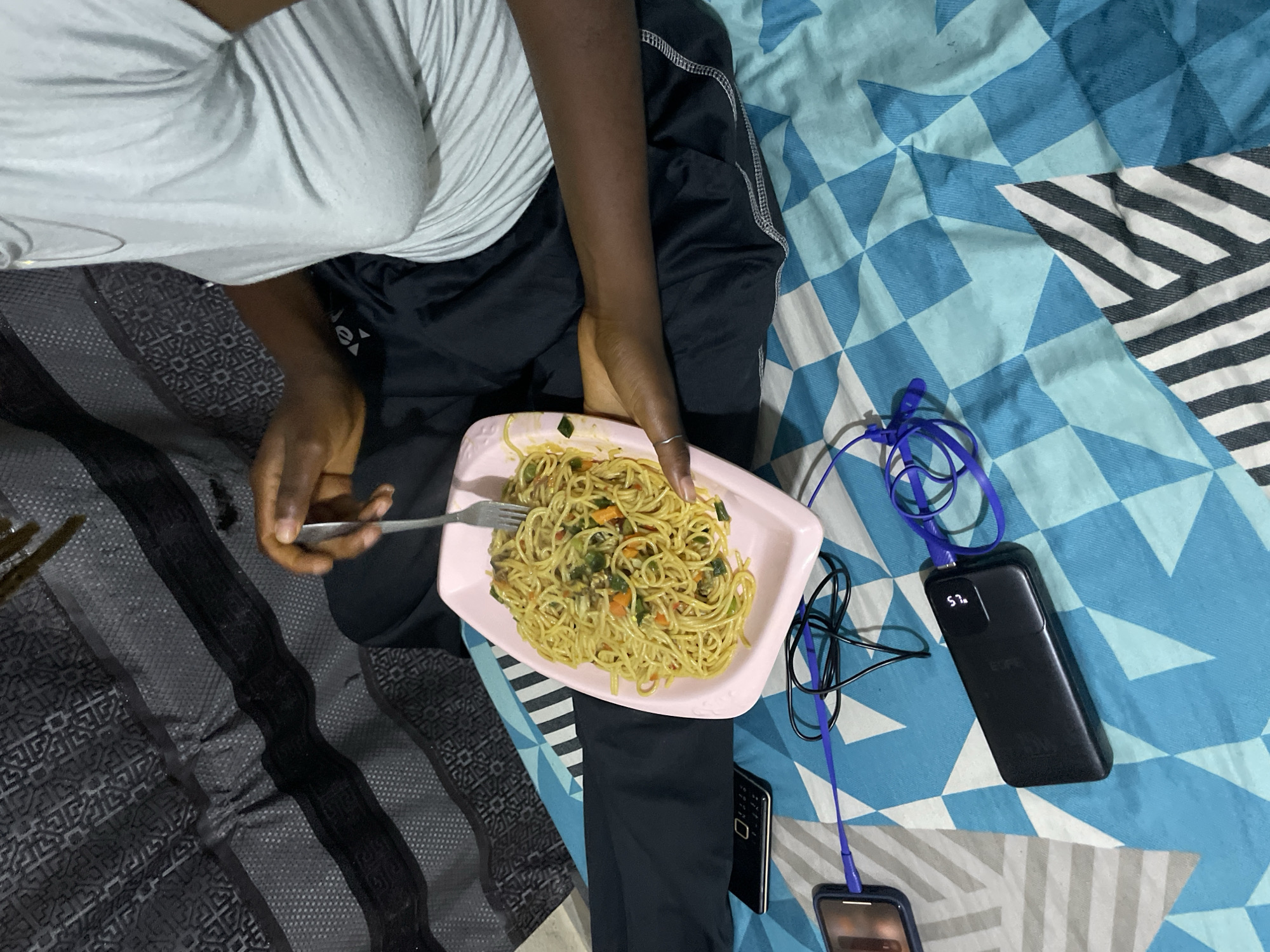 The image depicts a person, presumably of African descent based on skin tone, sitting on a blue and white patterned bedspread, likely indoors in an urban environment, possibly Rafin Par, Nigeria. They are wearing a light grey t-shirt and dark pants, with a silver necklace visible. The person is engaged in eating spaghetti from a pink rectangular plate using a fork. The spaghetti appears to contain vegetables such as carrots and green leafy vegetables.
On the bedspread to the left of the plate, a black flip phone is visible. Further to the left, a smartphone is connected by a blue charging cable to a black power bank, which is also connected to another set of blue headphones. The background shows a dark grey textured fabric, possibly a curtain or a bedsheet. The overall scene suggests a casual, indoor setting where the individual is relaxing and having a meal.