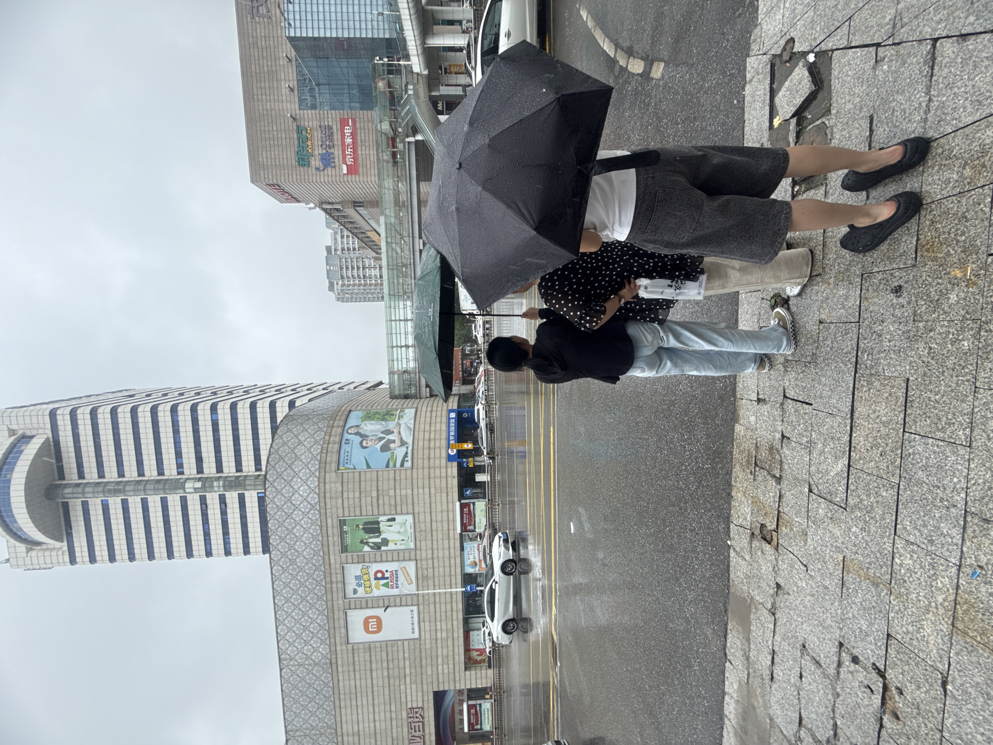 A group of three people are standing under umbrellas on a wet sidewalk in front of a large commercial building, likely a shopping mall, in Qinhuangdao, China. The building features a tall tower with a rounded top and numerous horizontal windows. The facade of the lower part of the building displays various advertisements, including logos for Xiaomi and "Russia". Across the street, a white sedan drives on the wet asphalt road, reflecting the overcast sky and surrounding buildings. The sidewalk is paved with granite tiles, some of which appear cracked or displaced. The weather is overcast and rainy, evidenced by the umbrellas and the wet surfaces.