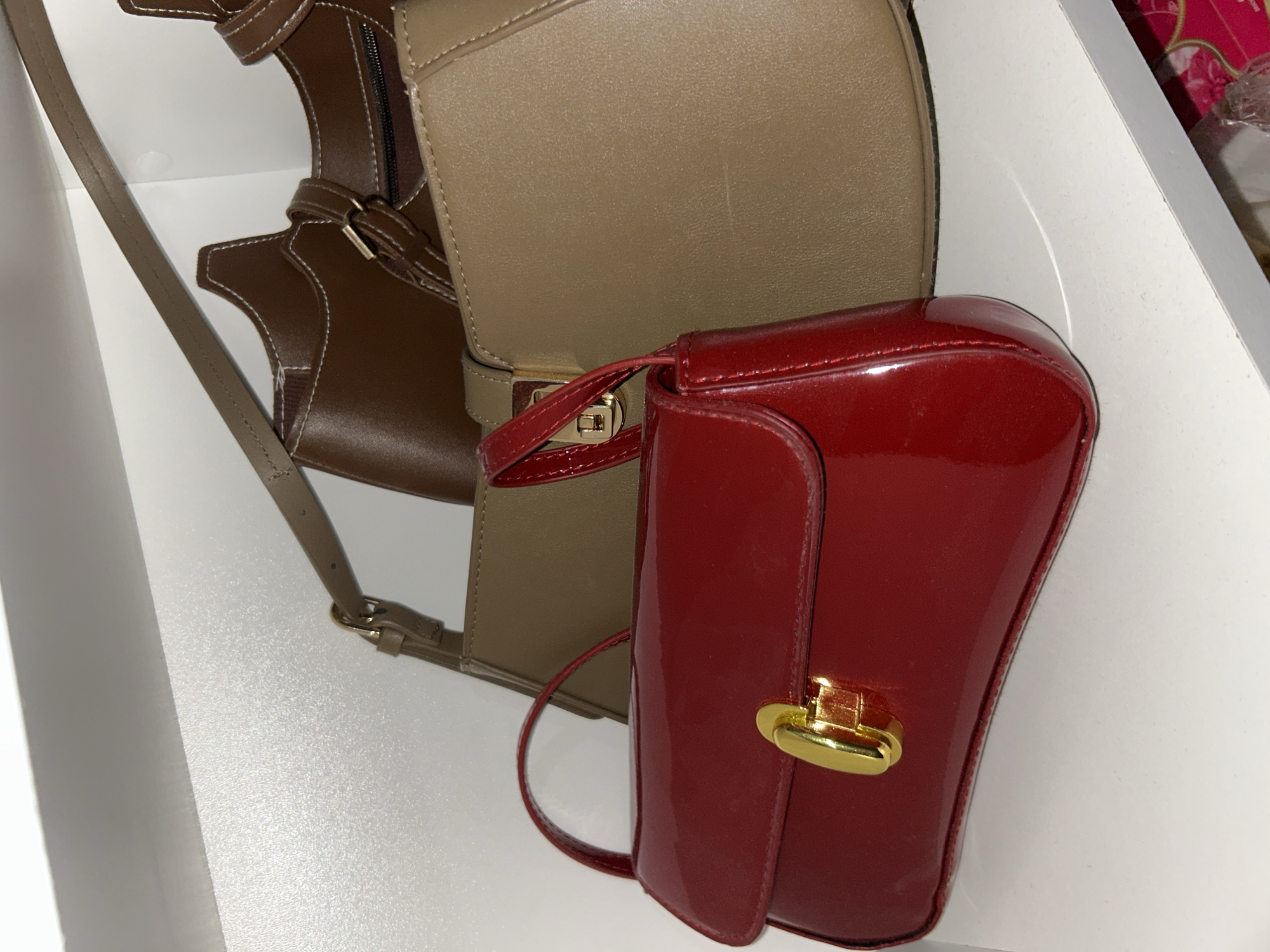 The image captures two handbags resting on a clean white surface, likely within a shelf or storage area. The background and surrounding surfaces are also uniformly white.
Prominently featured is a small, structured red handbag in the foreground, positioned to the right. It boasts a glossy, patent leather finish and is equipped with a distinct gold-toned turn-lock clasp on its front flap. A slender red strap is also visible, attached to the bag.
Behind and to the left of the red bag is a larger, light brown or tan handbag. This bag has a more matte, textured appearance, suggesting leather or faux leather. It features a flap closure secured by a gold-colored buckle or clasp. A darker brown section with visible white stitching and a zipper is attached or part of this handbag, partially extending from behind it. A long, light brown strap hangs from this bag.
No individuals are present, and no specific activity is occurring; the items appear to be stored. The lighting suggests an indoor setting. There are no visible brand names, logos, or other text. In the upper right corner, a small portion of a pink, patterned object, possibly a box or packaging, is just visible. There are no specific visual cues to indicate the stated location of Pleven, Bulgaria.