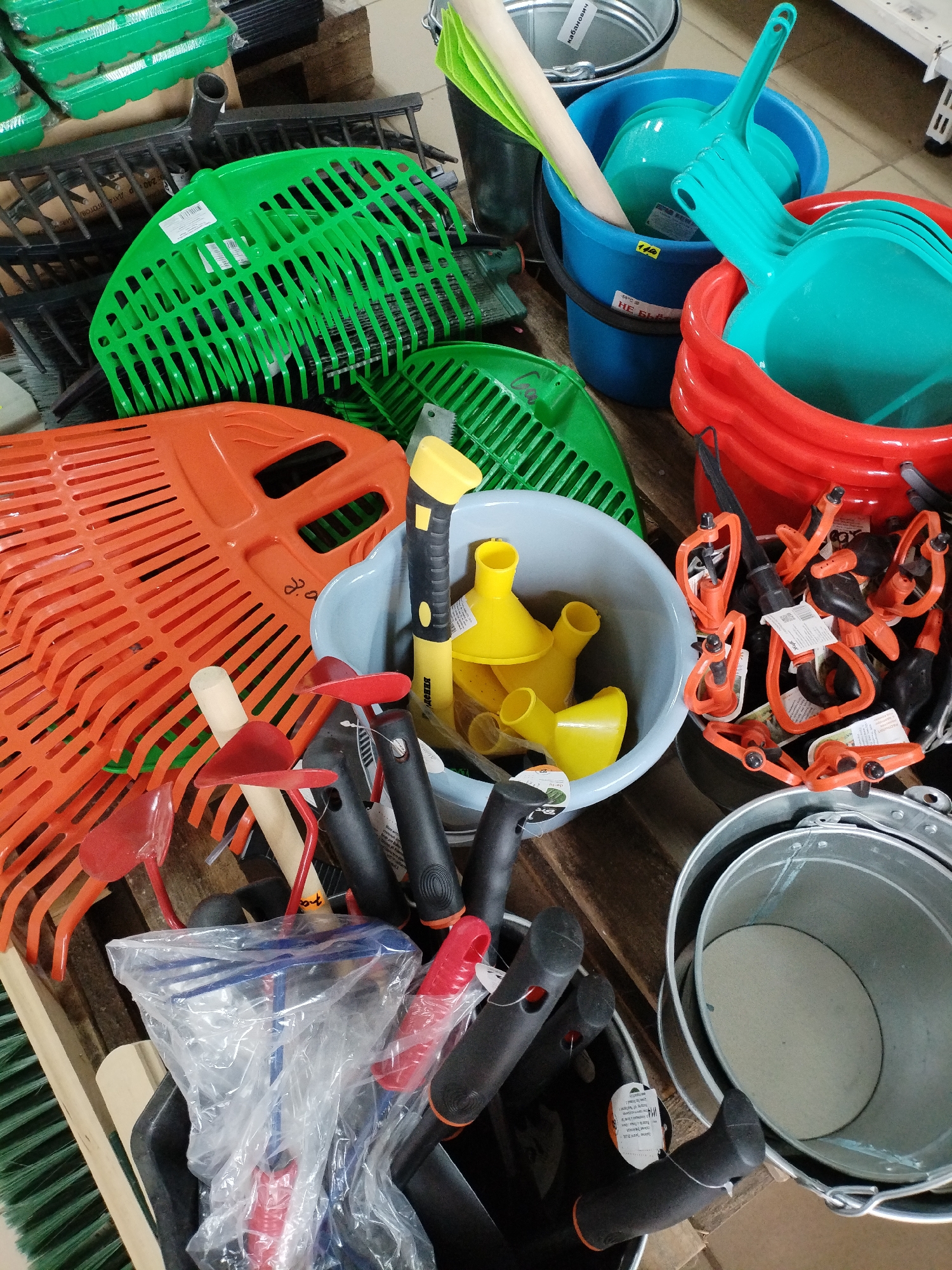 The image displays a close-up, top-down view of a retail display of gardening tools and buckets in Bialynichy, Belarus. The scene is cluttered with a variety of items, including several large, bright orange leaf rakes stacked on top of each other, a bright green fan rake, and smaller red hand trowels with wooden handles. Several blue and red plastic buckets are visible, some filled with turquoise plastic dustpans and others empty. A gray plastic basin contains several yellow funnels. A metal bucket is positioned in the foreground. The overall impression is of a disorganized but well-stocked display of gardening equipment.