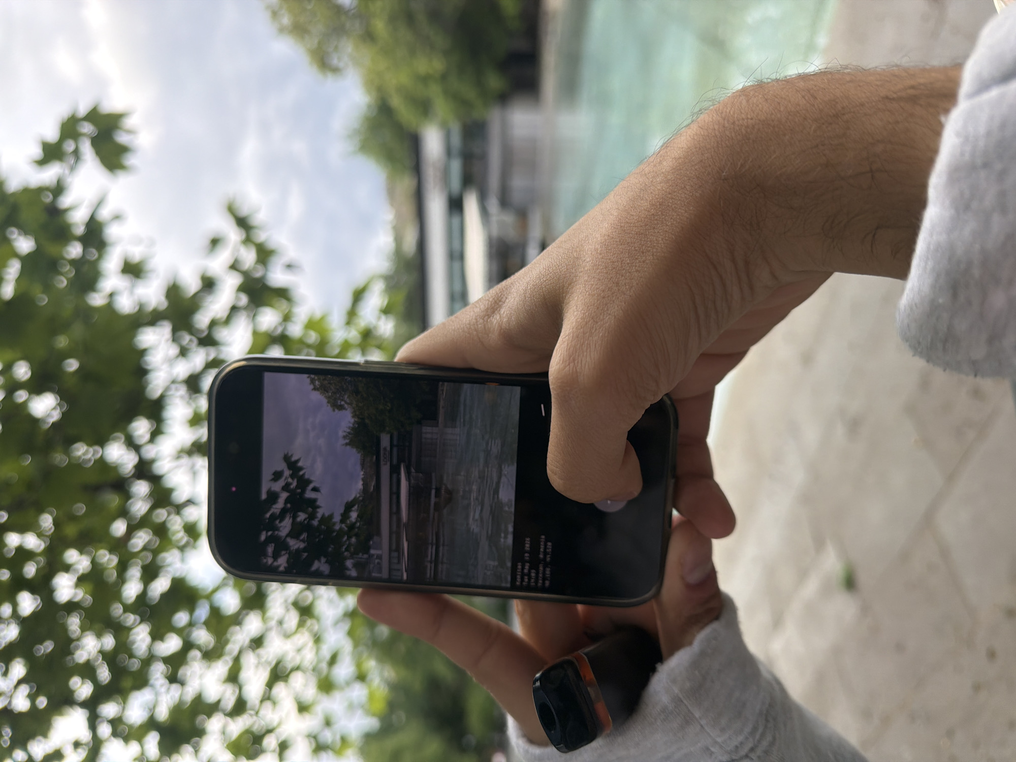 A person is holding a smartphone in front of them, displaying a photo of the Swan Lake in Yerevan, Armenia. The person's right hand is visible holding the phone, with a finger on the screen. Part of the person's left hand is visible holding a vaping device and wearing a gray sweatshirt. The foreground of the image is slightly out of focus and includes the treetops, while the background shows the Swan Lake, reflecting the surrounding buildings and trees. The sky is cloudy.