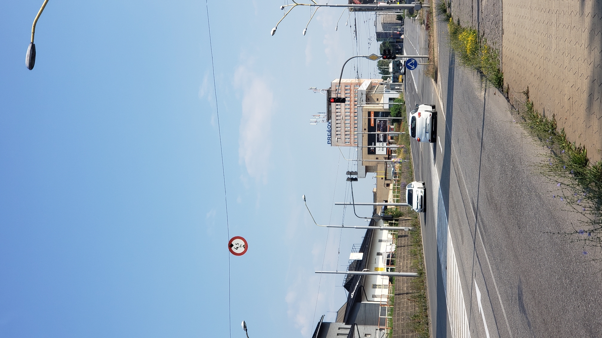 A bright, sunny daytime, outdoor, vertical, full-frame, medium shot captures a wide, paved road in Prešov, Slovakia. The sky is a clear, vibrant blue with a few wispy white clouds. The road is lined with sidewalks on both sides, with patches of green grass and small purple wildflowers growing along the edges.
In the foreground, the road is marked with white stripes, indicating a pedestrian crossing. Two white cars are visible on the road, one moving forward and another parked. Beyond the crossing, the road continues into the distance, lined with buildings of various sizes and architectural styles. A tall, multi-story building with a sign reading "PREŠOV" stands prominently in the background, adorned with telecommunication antennas on its roof. Streetlights are visible on both sides of the road, with traffic lights also present. A circular red and white traffic sign indicating a height restriction of 4.5 meters hangs suspended over the road from overhead wires. The scene is bustling with activity, with cars and a bus on the road and a few people visible in the distance near the buildings. The overall impression is that of a typical urban environment on a clear day.