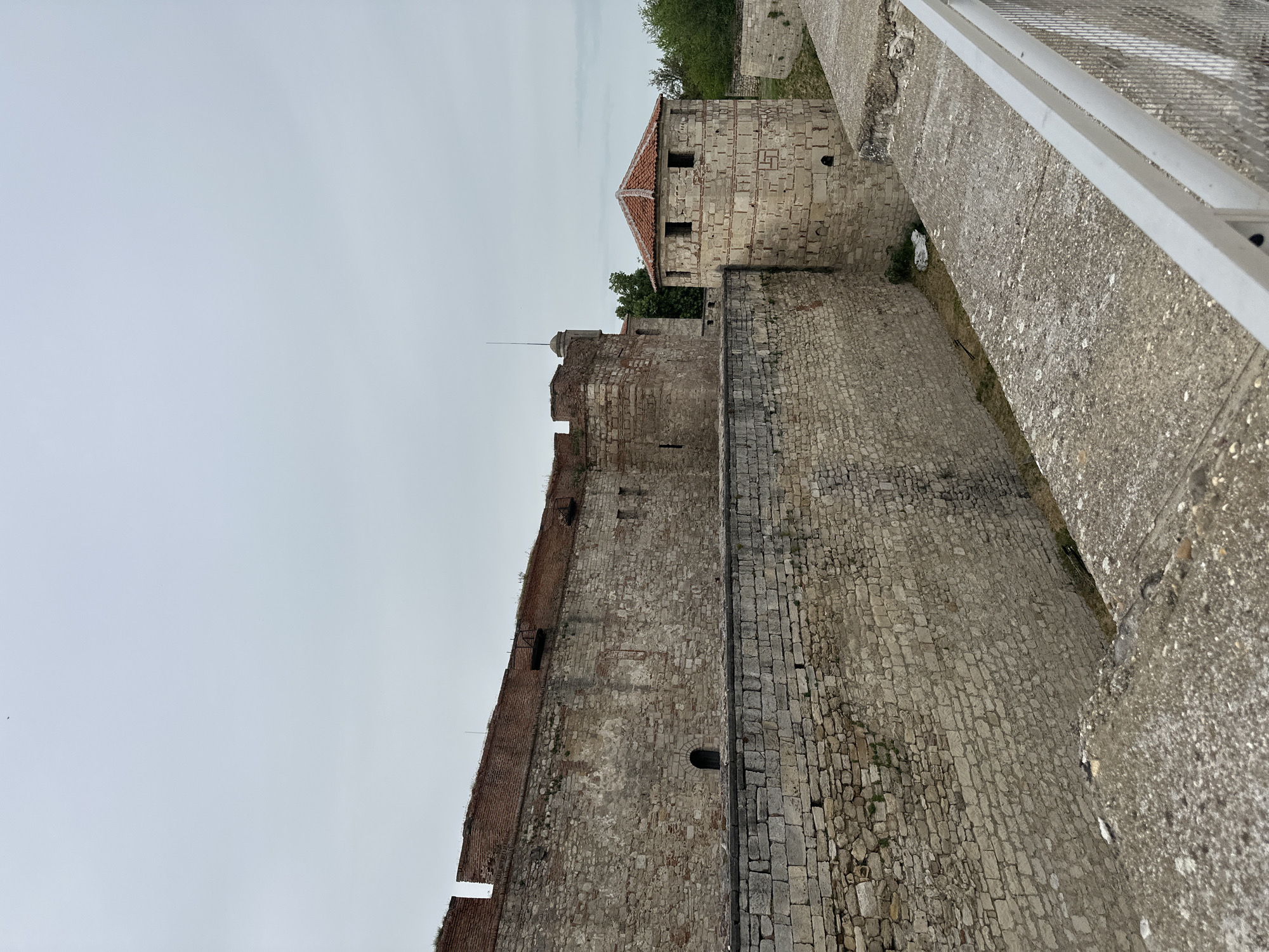 The image shows a section of the Vidin Fortress (Baba Vida), located in Vidin, Bulgaria. The fortress walls are constructed of stone and brick, with visible battlements and embrasures. One of the towers is visible with a red-tiled roof. There is a moat or trench running alongside the fortress wall. The image is taken from an elevated position, possibly from another part of the fortress or an adjacent structure. The sky is overcast. There are no people visible.