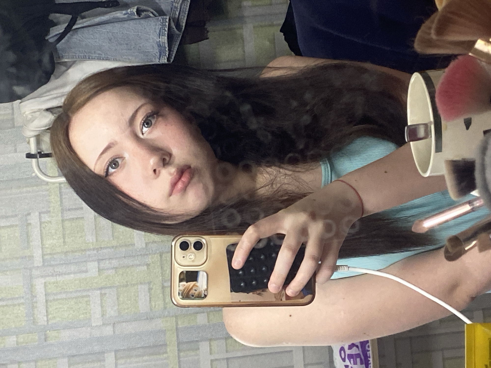 A young woman is taking a selfie in a mirror. She is holding a gold-colored smartphone with a black textured grip on the back. The phone has two rear cameras and a smaller square window that appears to display an image or sticker of a cartoon character. In her reflection, she is wearing a light blue top. Her long, dark brown hair is partly draped over her shoulder. She is looking directly at the camera with a neutral expression. Behind her, out of focus, are various objects: a piece of light-colored clothing, possibly denim, is visible in the upper-left corner. The location is not identifiable, and the city is unknown. Other objects, including makeup brushes, are also visible in the reflection, suggesting a personal space like a bedroom or bathroom.