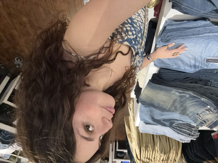 A woman with long, curly dark hair and fair skin is shown in a close-up shot. She wears a blue and white patterned top and several rings. Her arm rests on a stack of folded denim jeans in various shades of blue. A shelf with clothes is visible in the background. The image is rotated.