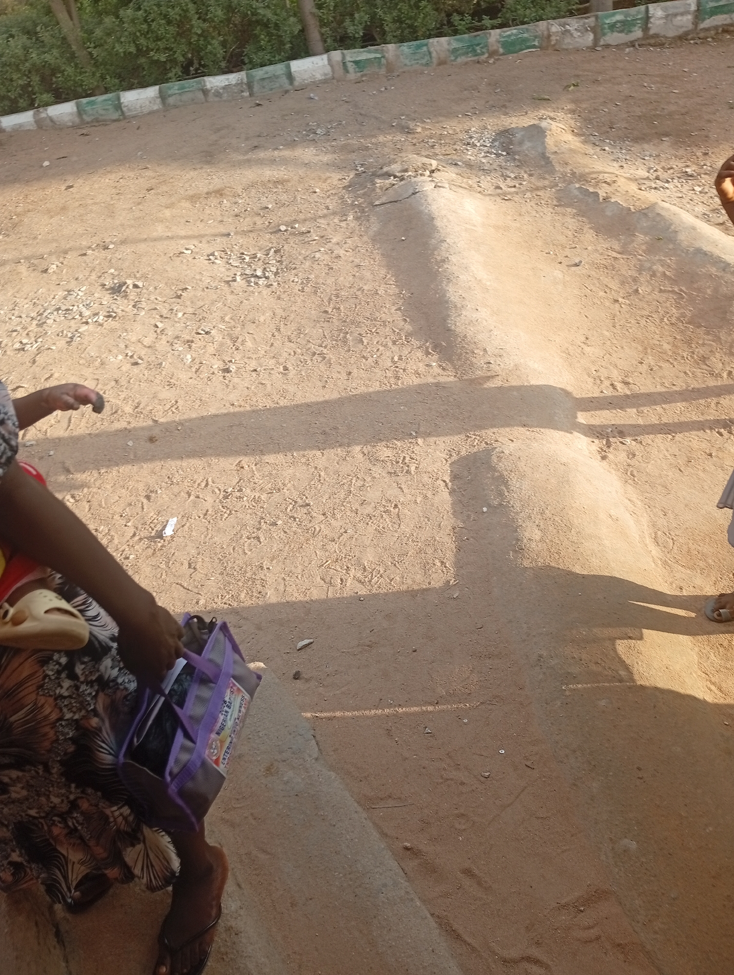 This media file depicts an outdoor scene with a dusty, sandy ground and vegetation in the background. Two individuals are partially visible, their presence suggested by their legs and hands. One person, on the left, is wearing a patterned skirt and holding a purple bag. Their hand is raised, and it appears they are either about to place something in or retrieve something from the bag. The other person, on the right, is only visible from the legs down, wearing sandals.
The ground is uneven, with some raised dirt mounds and scattered small stones. A concrete structure with green and white markings runs along the top of the frame, separating the sandy area from the vegetation. The lighting suggests it is daytime, with distinct shadows cast on the ground, indicating sunlight. The overall scene appears to be in a casual outdoor setting, possibly a pathway or an open area within a community.
The purple bag held by the person on the left has some visible text on it. The text includes "THE NIGERIAN BAPTIST" and "ENTER...", suggesting it might be a bag associated with the Nigerian Baptist Convention. No specific activity or event is clearly identifiable, but the gesture of holding the bag implies it is in use. The location context is provided as Jalingo, Nigeria.