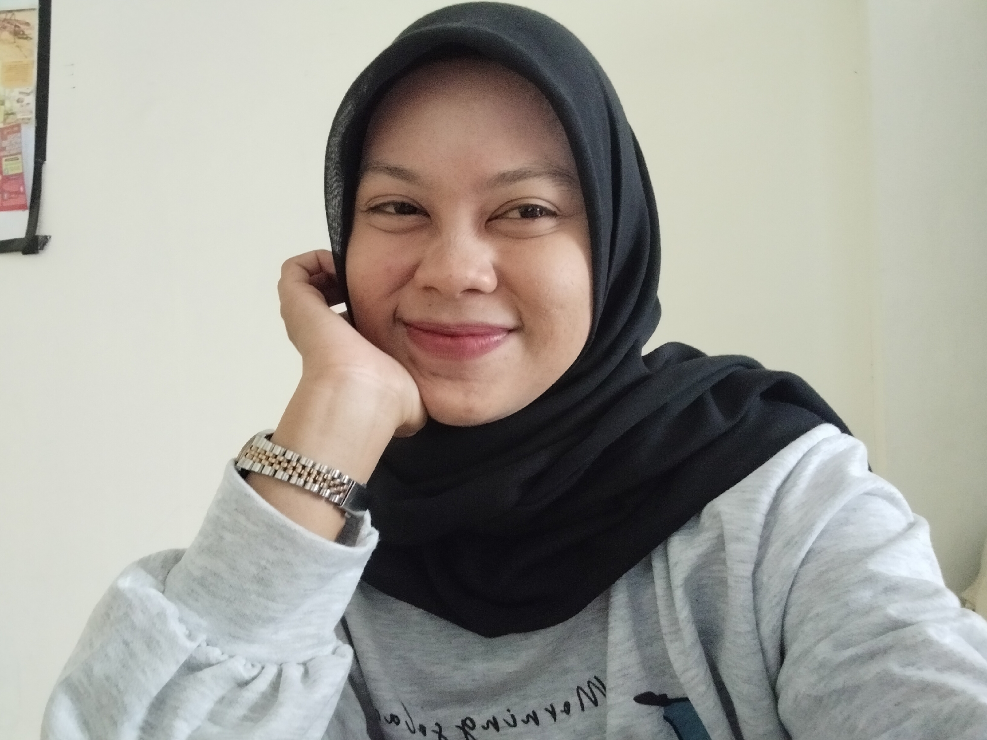 A young woman wearing a black hijab and a grey long-sleeved shirt is looking at the camera with a slight smile. She has her right hand resting on her chin, and she is wearing a watch on her left wrist. The background is a plain white wall. There is a framed picture on the wall to the left of the woman. The scene appears to be indoors. The woman's clothing suggests a warm climate, consistent with the location of Julimeunasah Teungoh, Indonesia.