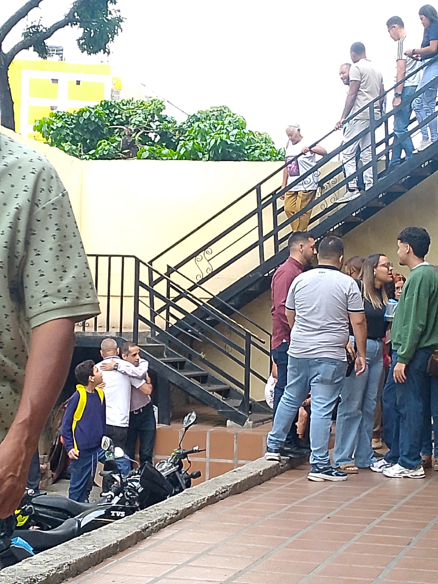The image captures an outdoor scene in Caracas, Venezuela, featuring a group of people on and around a set of exterior stairs.
People: The photograph shows a diverse group of individuals of various ages and genders. On the stairs, some people are ascending, including an elderly woman. Near the base of the stairs, a group is gathered in conversation. There is an embrace happening between two men, with a young boy nearby.
Environment: The backdrop includes a yellow building with white trim, partially obscured by green trees. The stairway has black metal railings. The ground is paved with orange-brown rectangular tiles.
Objects: Two motorcycles are parked next to the curb.
Actions/Interactions: People are ascending the stairs, conversing in a group, and embracing each other.
Subject: The focus appears to be on the interactions and gatherings occurring around the stairway.