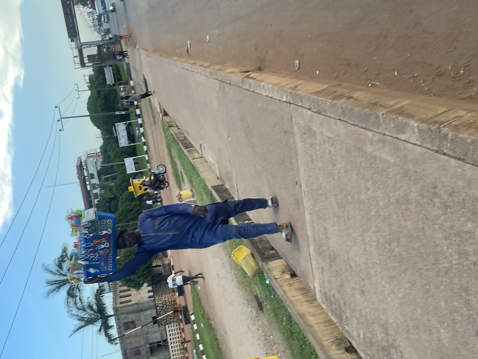 A man in a navy blue traditional outfit walks along a concrete sidewalk. He is carrying a large blue plastic basket filled with assorted items on his head. The basket contains numerous keychains and other small goods. He is wearing sandals on his feet. Behind him, a yellow plastic bucket lies on its side on the grass next to the sidewalk. Further in the background, there is a street with moving vehicles and a large gate structure. The scene appears to be in an urban environment, potentially a marketplace or a busy street in the city of Jos, Nigeria, as indicated by the presence of palm trees and architectural styles common in the region. The sky is partly cloudy.