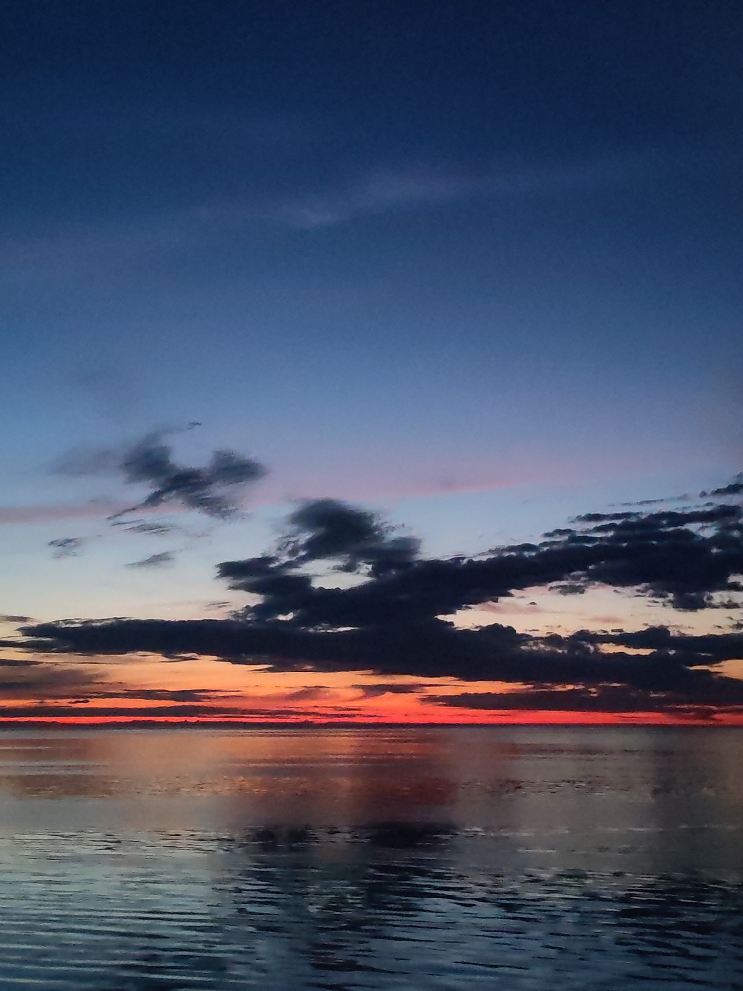 The image shows a body of water under a dramatic sunset sky. The sky transitions from deep blue at the top to shades of orange and red near the horizon, with dark clouds scattered across the middle of the frame. The water reflects the colors of the sky, creating a mirrored effect. The location of the image is not visible. There are no people, subjects, objects, actions, or interactions visible in this image.