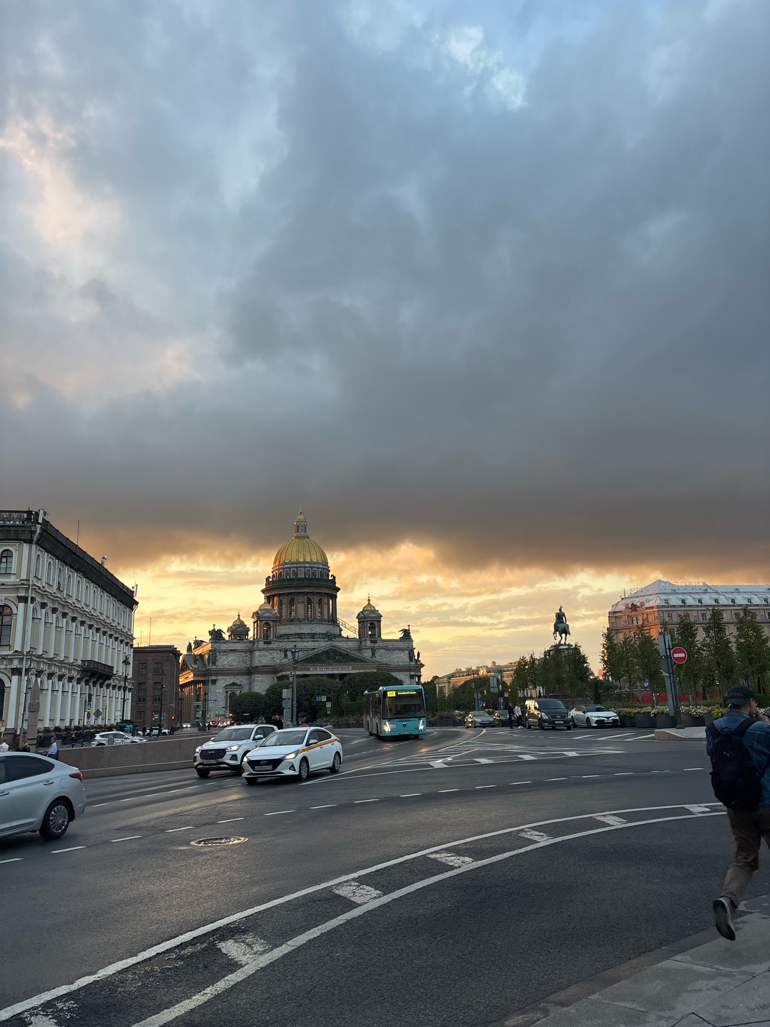 This is a photograph of St. Isaac's Cathedral in Saint Petersburg, Russia, during sunset. The sky is filled with dramatic clouds, with the setting sun casting a warm, golden light on the horizon. The cathedral, with its iconic golden dome, is the central focus of the image. In the foreground, a street scene unfolds with cars, a bus, and pedestrians. The architecture of the surrounding buildings, typical of Saint Petersburg, is also visible. The overall mood is atmospheric, capturing the beauty of the city at dusk.