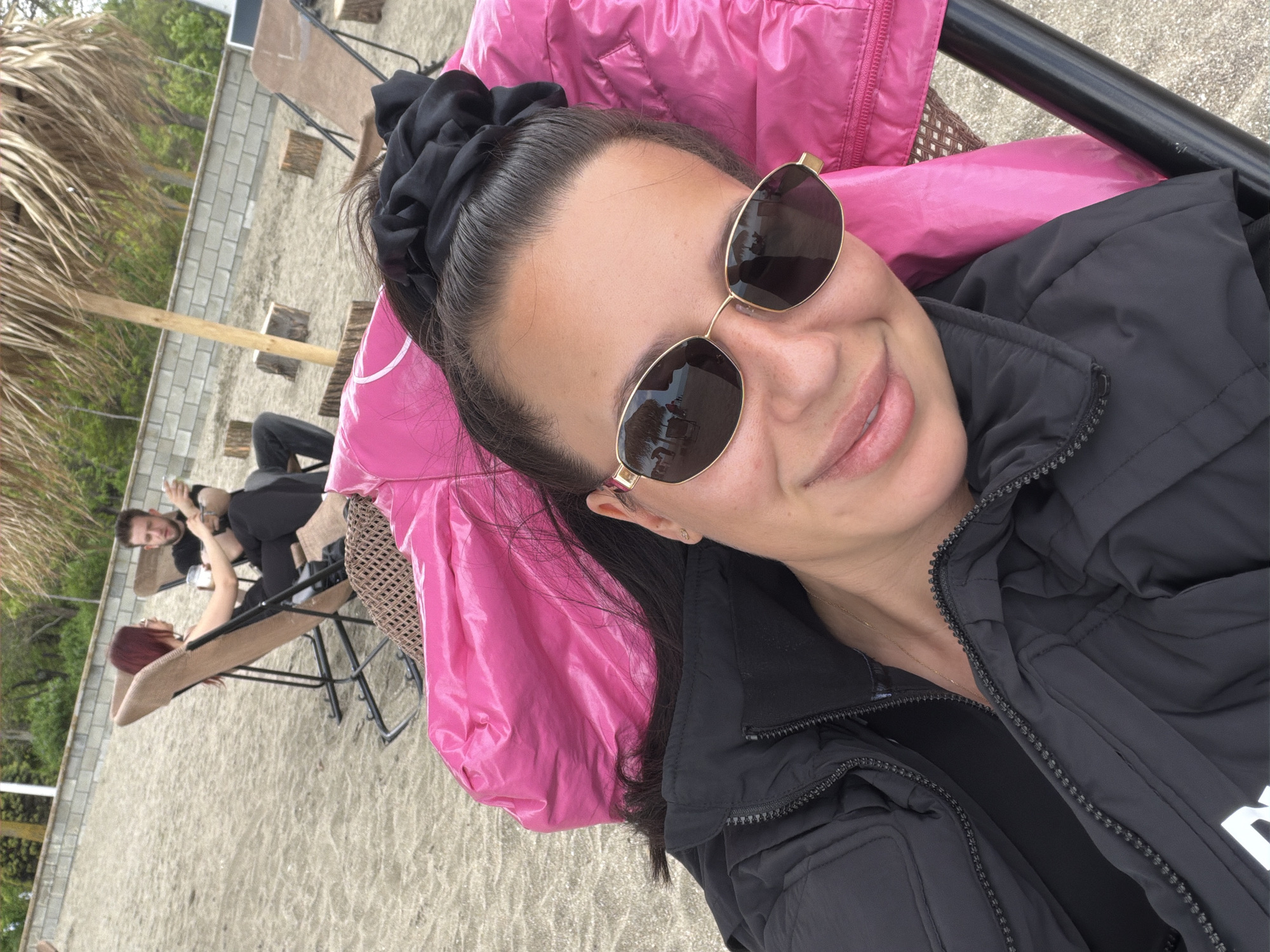 A selfie photo taken on the beach in Burgas, Bulgaria. A woman with dark hair pulled up in a black scrunchie and sunglasses is lying on a lounge chair covered with a pink jacket. She is wearing a black jacket. In the background, two people are visible on lounge chairs near a palm tree and a white brick wall. The ground is sandy.