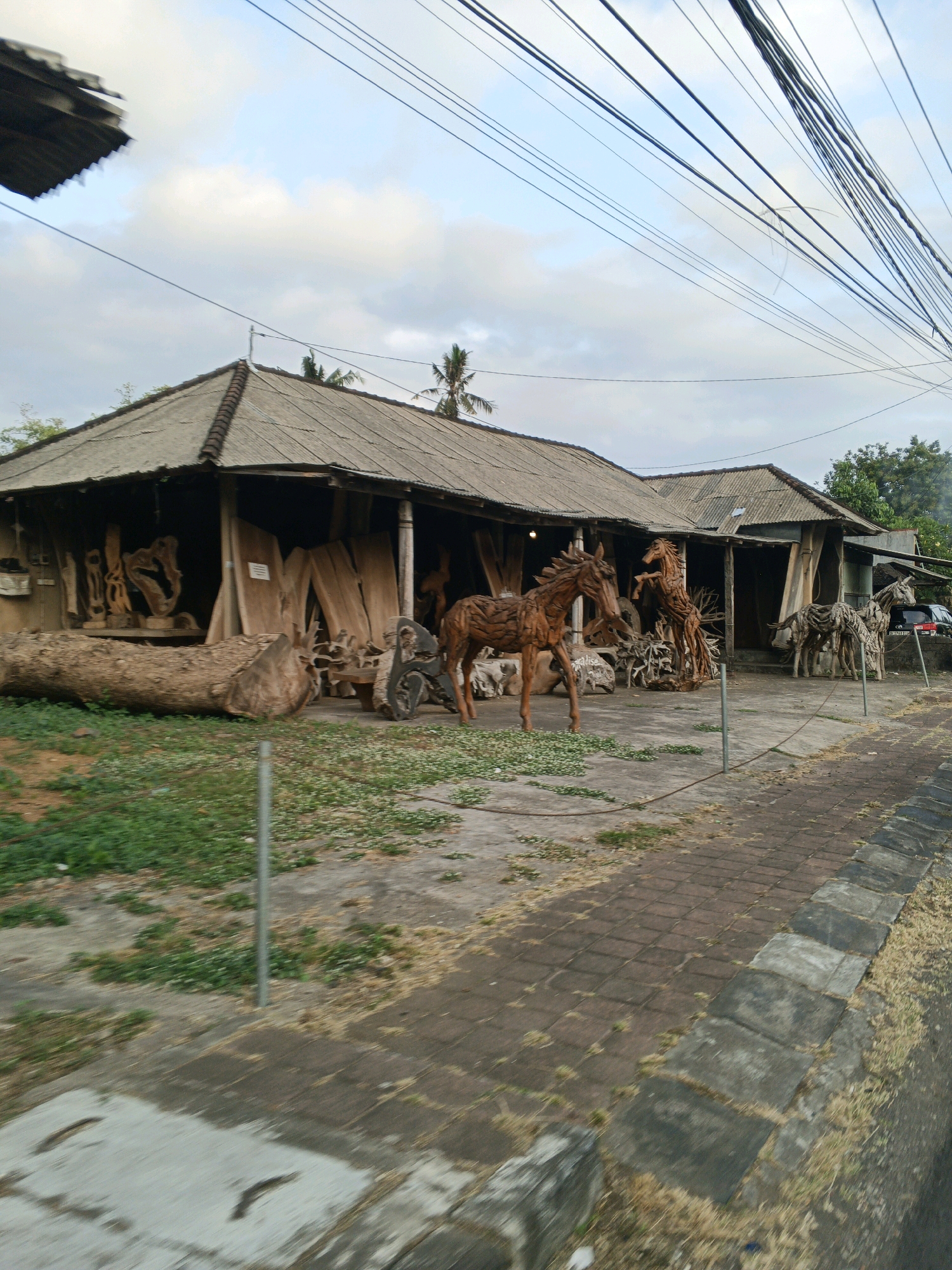 The image depicts an outdoor art gallery or workshop in Sukawati, Indonesia, showcasing various wooden sculptures. The primary subjects are several intricately carved wooden horses, arranged in the foreground and mid-ground of the scene. One horse, in the center, is standing with its head raised. To its right, another horse appears to be rearing. Further to the right, there are more horse sculptures, one appearing as a herd, and another in a galloping stance.
The structures in the background are open-air stalls with corrugated metal roofs, typical of street-side businesses in the region. These stalls display a variety of raw lumber and finished wood carvings, suggesting a woodworking craft. Large logs are also visible, including a prominent one in the left foreground. The ground in front of the stalls is a mix of concrete, dirt, and scattered green vegetation. A paved walkway, made of reddish-brown bricks and bordered by concrete slabs, runs along the right side of the image, indicating a public access area. Overhead, a dense network of electrical wires crisscrosses the sky, a common sight in Indonesian cities. A dark-colored vehicle is parked behind the structures on the right. The lighting suggests it is either early morning or late afternoon, with soft, diffused light and overcast skies. The overall scene conveys a sense of a bustling artisan marketplace.