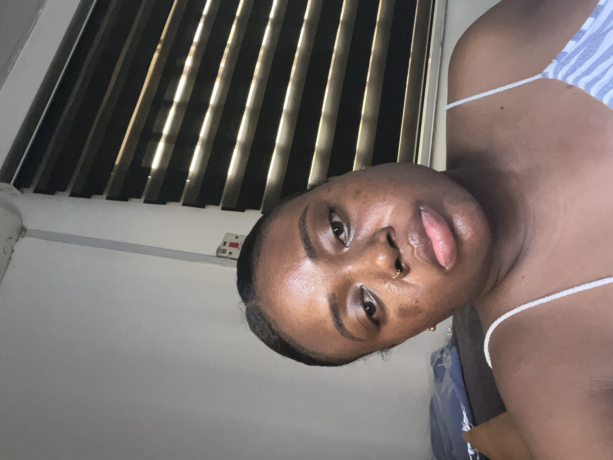 A close-up portrait of a Black woman indoors. She is wearing a white strap top with a blue and white striped pattern on the top. Her skin has visible pores and some blemishes. She has a gold nose ring in her left nostril and a gold earring is partially visible. The background features closed window blinds that are dark with bright light filtering through. A white wall is visible on the left, with an electrical outlet near the top. Part of a blue garment can be seen behind her shoulder. The location and city is Abuja, Nigeria.