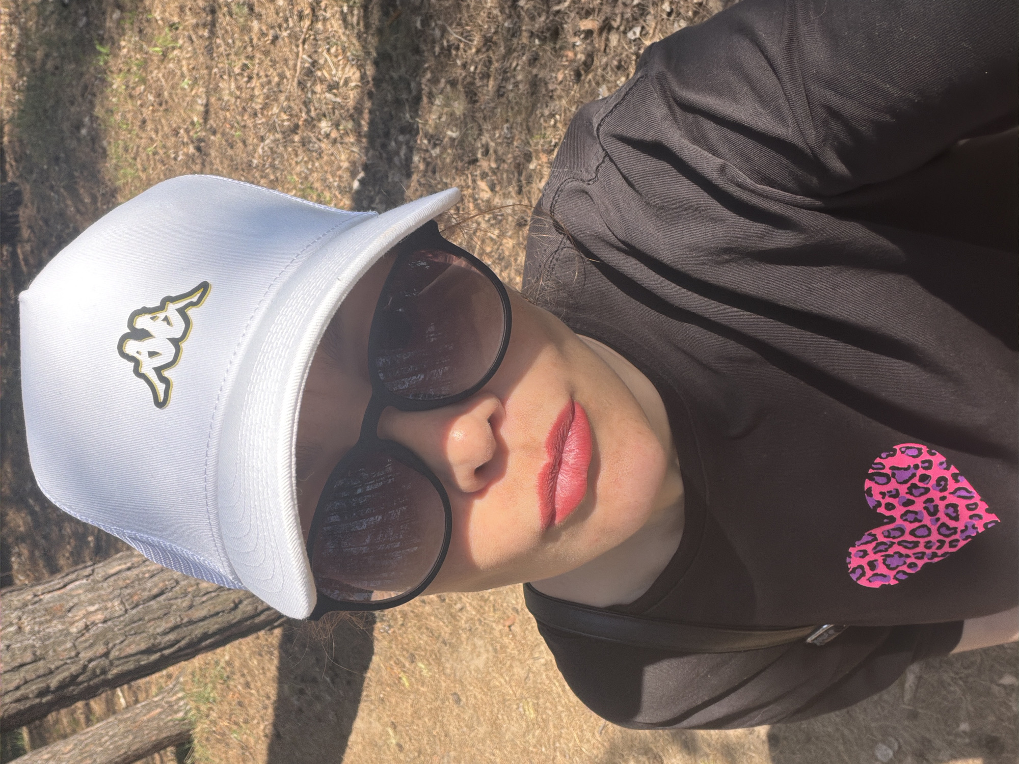 A woman is posing outdoors, possibly on a hill or near trees, as suggested by the dry vegetation and tree limbs visible in the background. She is wearing a white Kappa-branded baseball cap, round sunglasses, and a black t-shirt with a pink and purple leopard-print heart design. Her lips are red. The location is Chaikovsky, Russia.