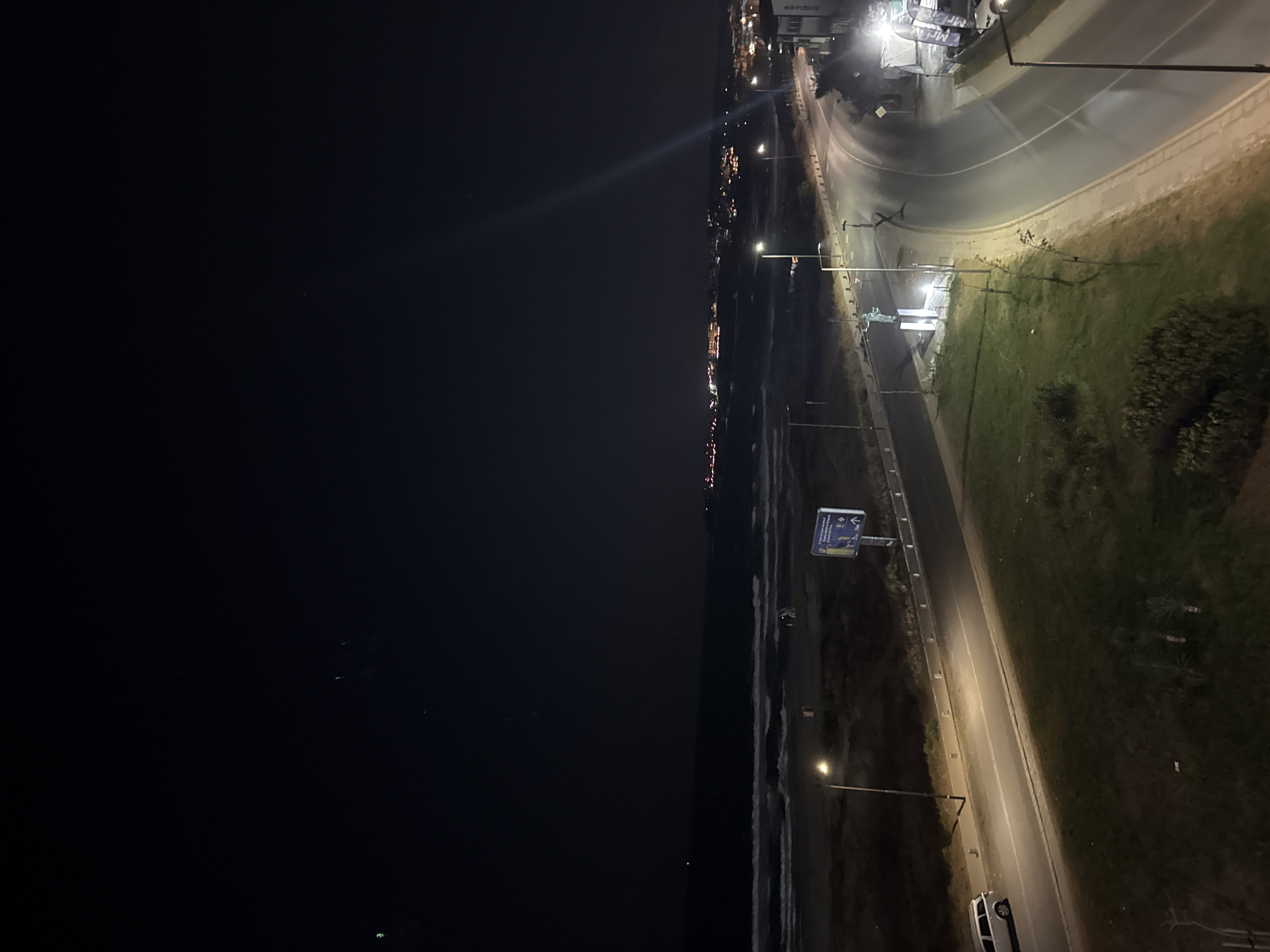 This is a nighttime, wide-angle, aerial view of a coastal area in Sozopol, Bulgaria. The scene captures the Black Sea, a beach, a road, and a distant town illuminated by lights.
The sea is dark with visible waves, suggesting it is choppy. The beach area appears sparsely populated, with a few scattered beach chairs or umbrellas visible. A lamppost stands near the beach.
A road curves through the foreground and midground, illuminated by streetlights. There is a white station wagon parked on the side of the road. Adjacent to the road is a grassy area with some small trees or bushes. Further along the road, a statue of a person with outstretched arms stands on a pedestal, illuminated by spotlights. There are also several signs and what appear to be advertising banners near the statue.
In the background, a town stretches along the coastline. Numerous lights from buildings and streetlights create a warm glow against the dark sky, indicating a populated area. A faint beam of light, possibly from a spotlight or searchlight, extends upwards into the sky from the town. The sky itself is dark, with a few faint lights visible, which could be stars or distant aircraft.