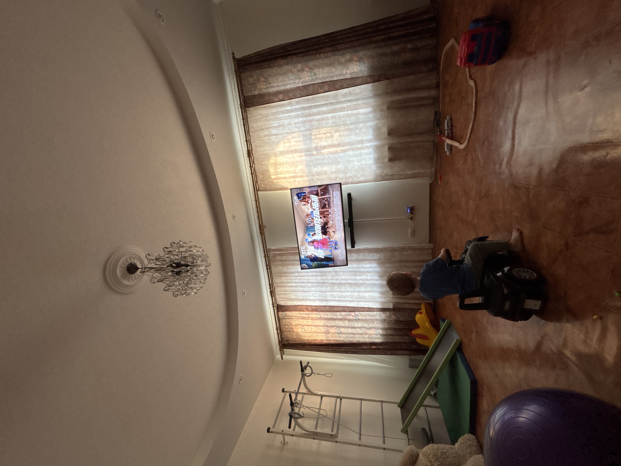 The image shows an interior scene, possibly a child's playroom, in Ферма, Russia. The room is characterized by a beige tile floor and a complex ceiling with a large chandelier. In the center of the image, a television is mounted on the wall, displaying a children's show. A window behind the television is partially covered by patterned brown curtains.
A young child wearing a blue shirt is riding a black toy car. A slide is positioned adjacent to the toy car, leading down from a small elevated platform. A set of gymnastic wall bars are in the background near the slide, and a large purple exercise ball sits on the floor in the foreground. There are also other toys scattered around the room, including a blue and red toy and a yellow toy near the slide.