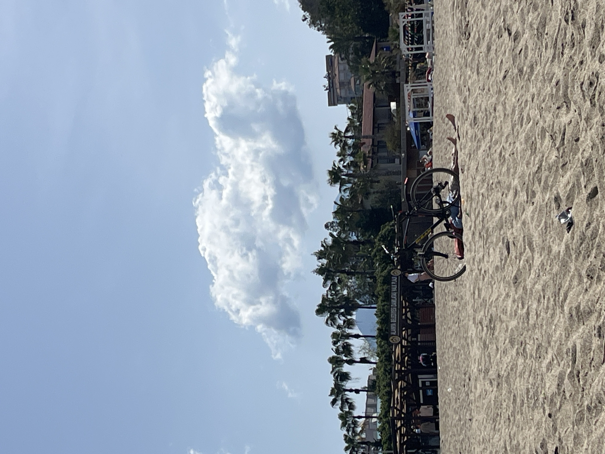 The scene is a beach in Alanya, Turkey, specifically on Damlataş Beach (indicated by a sign). The beach has light-colored sand. A bicycle is parked on the beach. A person is lying on the beach in the background. Palm trees line the edge of the beach near buildings. There are white clouds in a blue sky.