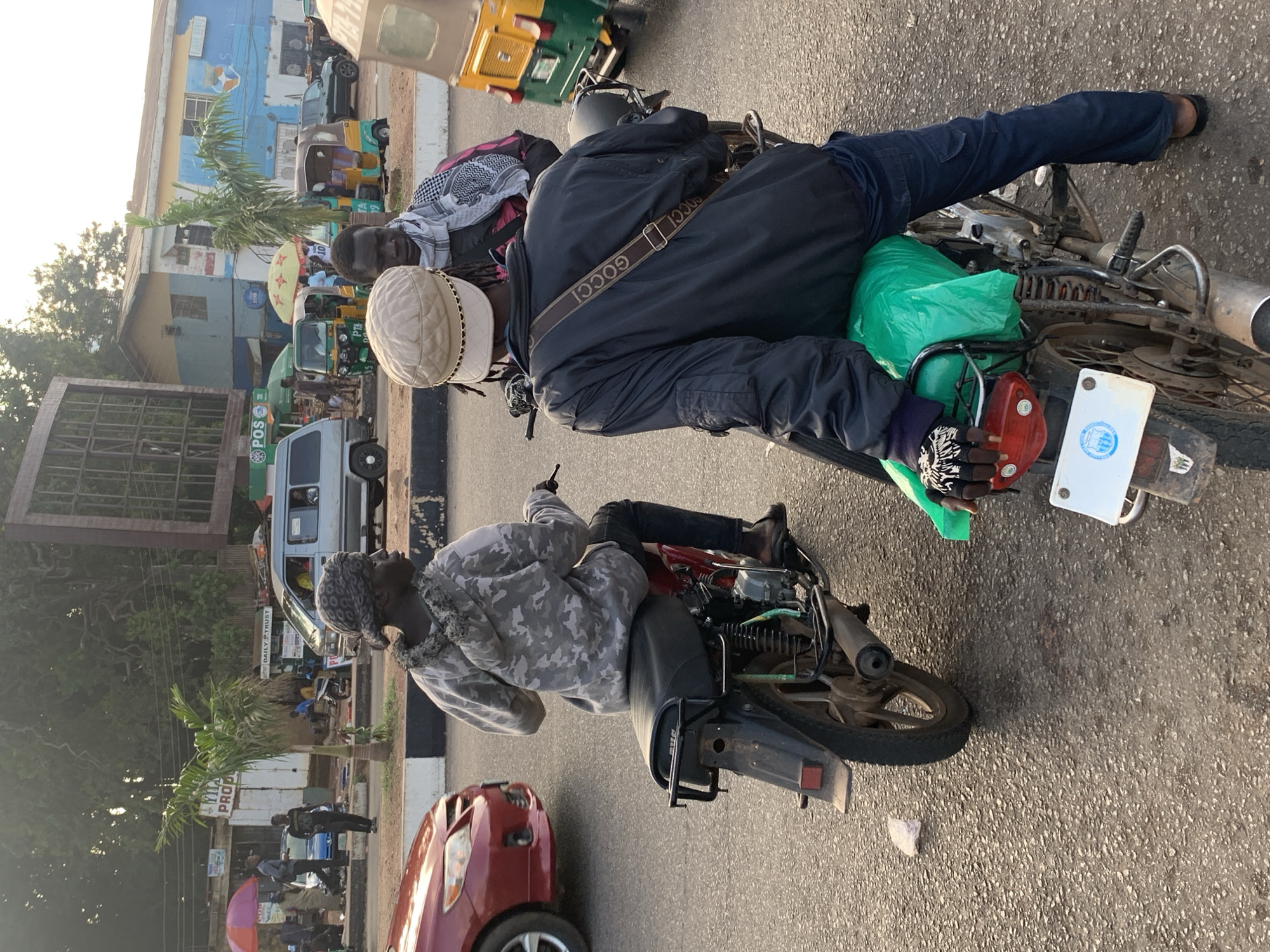 The media file captures a scene in Barakin Lerawi, Nigeria, likely during daylight. The foreground features two individuals on a motorcycle. The person in the front is wearing a light-colored patterned jacket and a headscarf, with their head turned away from the camera. The person behind them is wearing a dark jacket, dark pants, and a beige cap, and appears to be the driver. A green plastic bag is visible on the back of the motorcycle, along with other items. A strap with the word "GUCCI" is visible across the driver's jacket. The motorcycle itself is a common model, with visible suspension and exhaust.
In the background, a bustling street scene unfolds. Several other motorcycles and a tricycle (also known as an 'okada' or 'bajaj' in Nigeria) are present, suggesting a busy transportation hub. Buildings with various signage are visible, including one with blue and yellow paint and what appears to be a logo. A red car is parked in the lower right corner of the frame. The overall impression is of a typical street scene in a Nigerian town, with people and vehicles navigating the area. The lighting suggests it's either morning or late afternoon.
