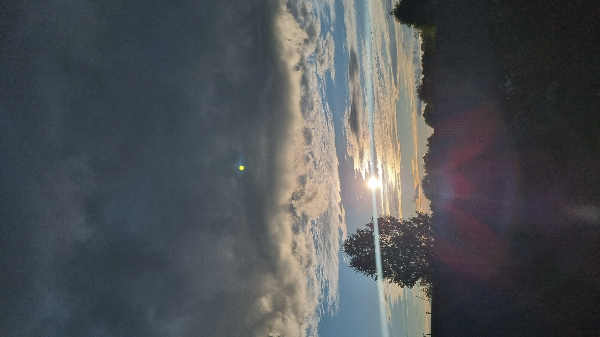 This image captures a rural landscape at sunset, likely in Vysokoe, Russia, given the provided context. The sky is a dominant feature, with a dramatic contrast between dark, heavy clouds in the upper portion and lighter, textured clouds illuminated by the setting sun in the mid-ground. The sun itself is a bright, horizontal flare low in the sky, casting a warm glow and creating lens flare artifacts that streak across the lower half of the image. A single, silhouetted tree stands prominently on the left, with a line of darker trees forming a horizon in the distance. The foreground is a dark, indistinct field or ground, heavily impacted by the low-angle light and lens flare. There are no people or discernible actions taking place, presenting a static natural scene.