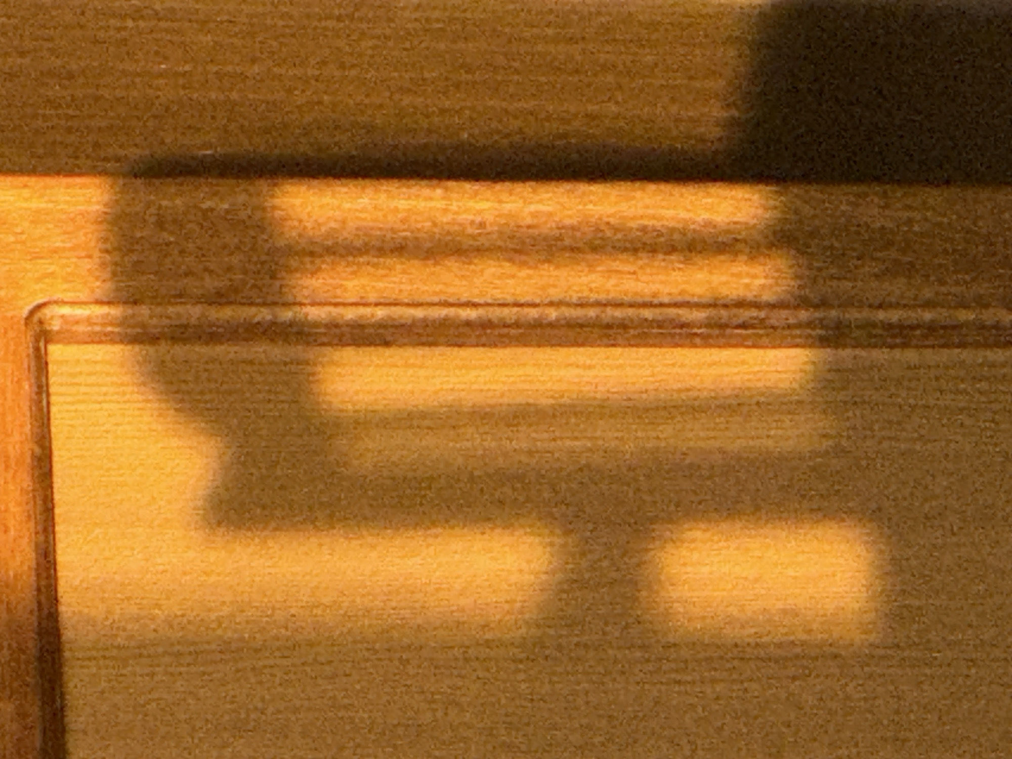 The image shows a close-up of a wooden surface, likely a door or wall, with shadows cast upon it. The wood grain is visible, with a warm, orange-toned light illuminating parts of the surface and casting distinct shadows. The shadows form abstract shapes, resembling a hand or fingers reaching out and some vertical lines, possibly from a fence or blinds. The lighting suggests a warm, possibly late afternoon or early morning sun. There are no people or specific identifiable objects in the scene beyond the wooden surface and the shadows. The environment is an indoor setting with a wooden interior surface.