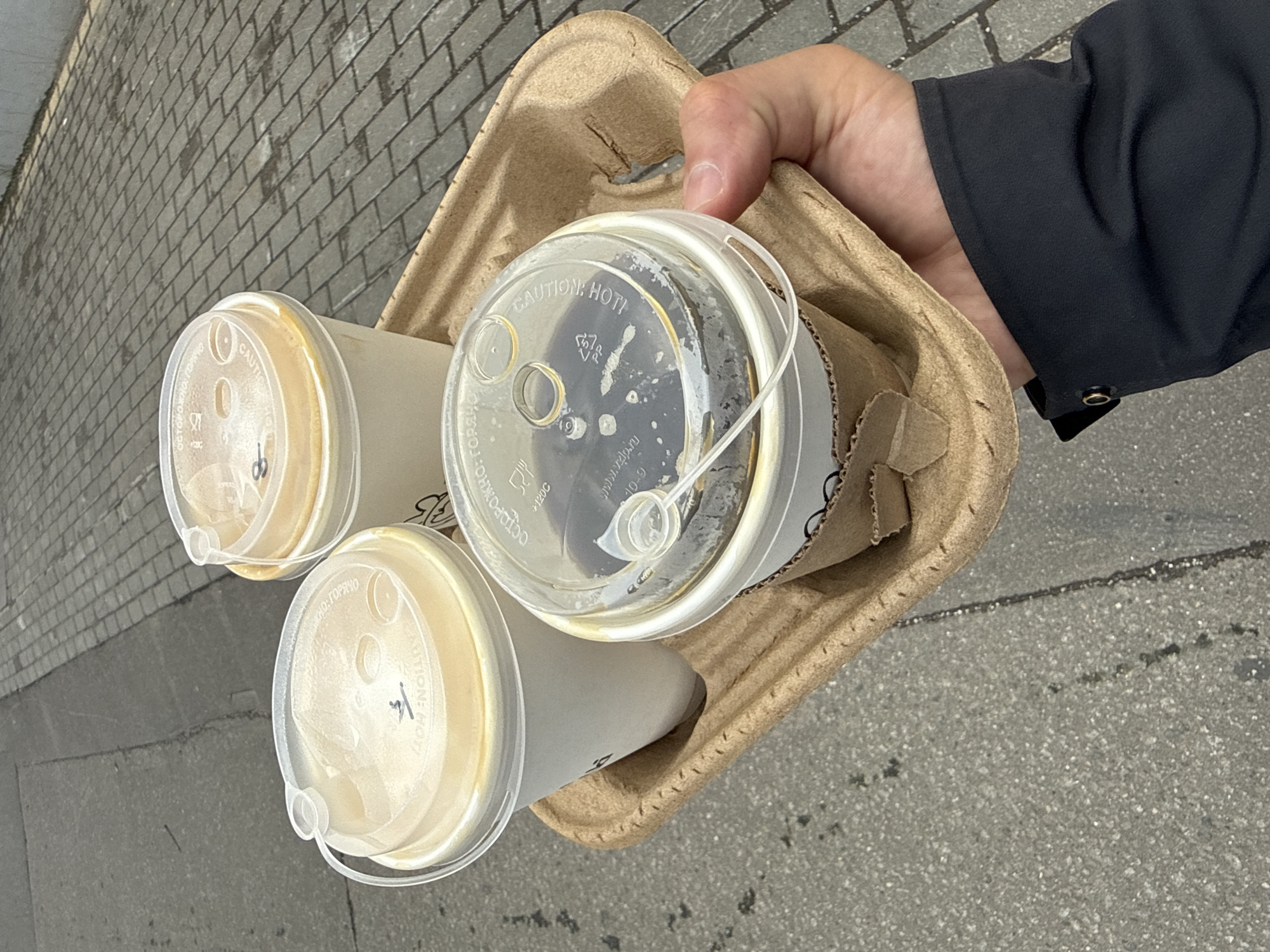 A person's hand, wearing a dark-colored jacket with a button on the cuff, holds a cardboard coffee carrier containing three cups of coffee with clear plastic lids. The tops of two cups have a light tan beverage inside, while the third, in the center, appears to contain a darker liquid. Cyrillic writing is visible on the lids, along with the words "CAUTION: HOT!" The carrier is brown, and the scene is shot at an angle, with the ground visible in the background. The ground has a section of brick paving stones, suggesting an urban environment, possibly in Moscow based on the Cyrillic text on the lids.