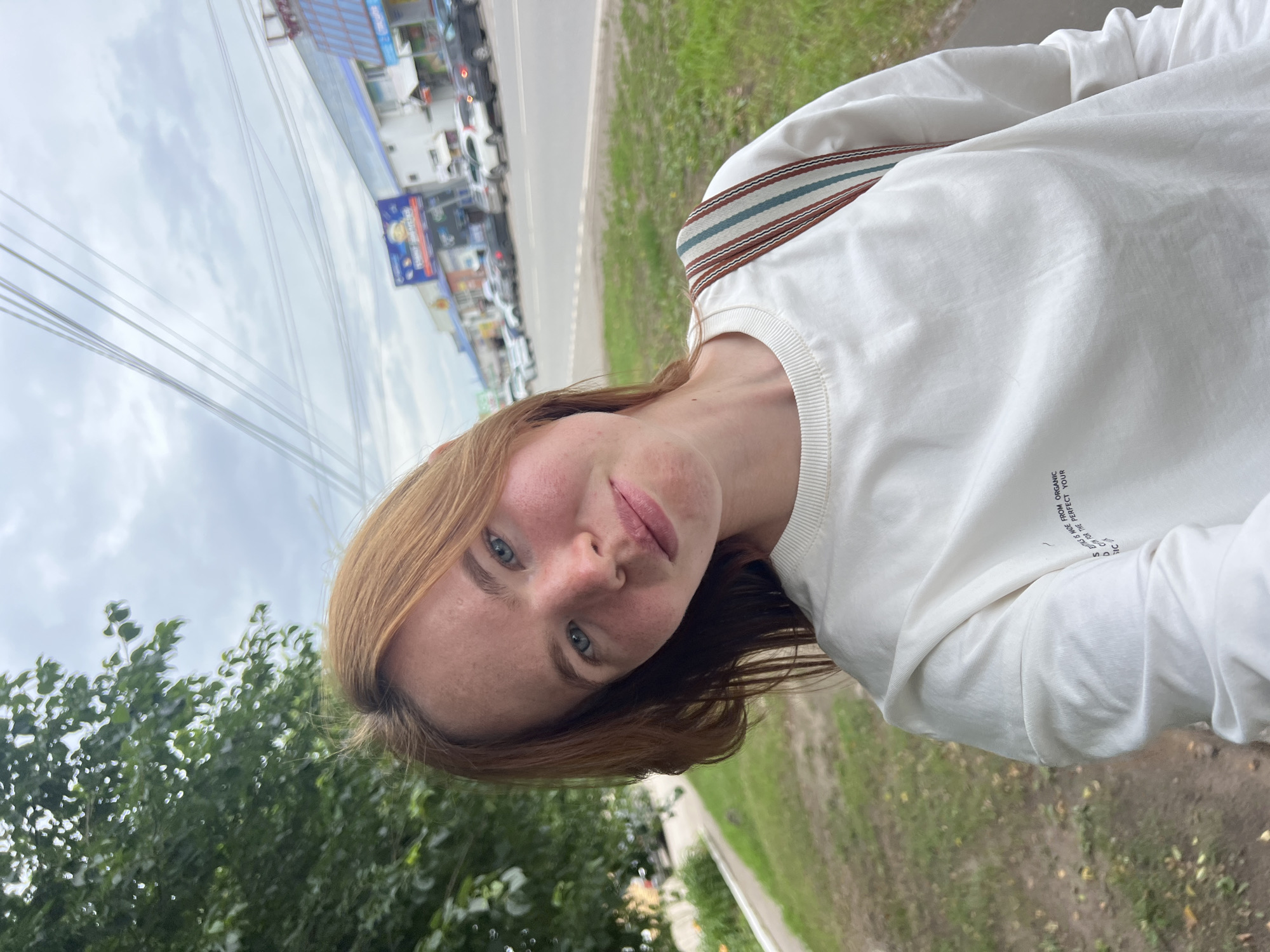 The image is a portrait of a young woman with reddish-brown hair and blue eyes, smiling at the camera. She is wearing a white long-sleeved shirt with a striped detail on the shoulder. The image is taken outdoors, with a street lined with buildings and cars visible in the background. Green trees are also present in the foreground, and the sky appears cloudy. The woman's face is slightly flushed, and she has freckles. There is some text visible on her shirt, though it is partially obscured and difficult to read clearly. The overall impression is casual and relaxed. The lighting suggests it is daytime, possibly overcast. The location is Chaikovsky, Russia.