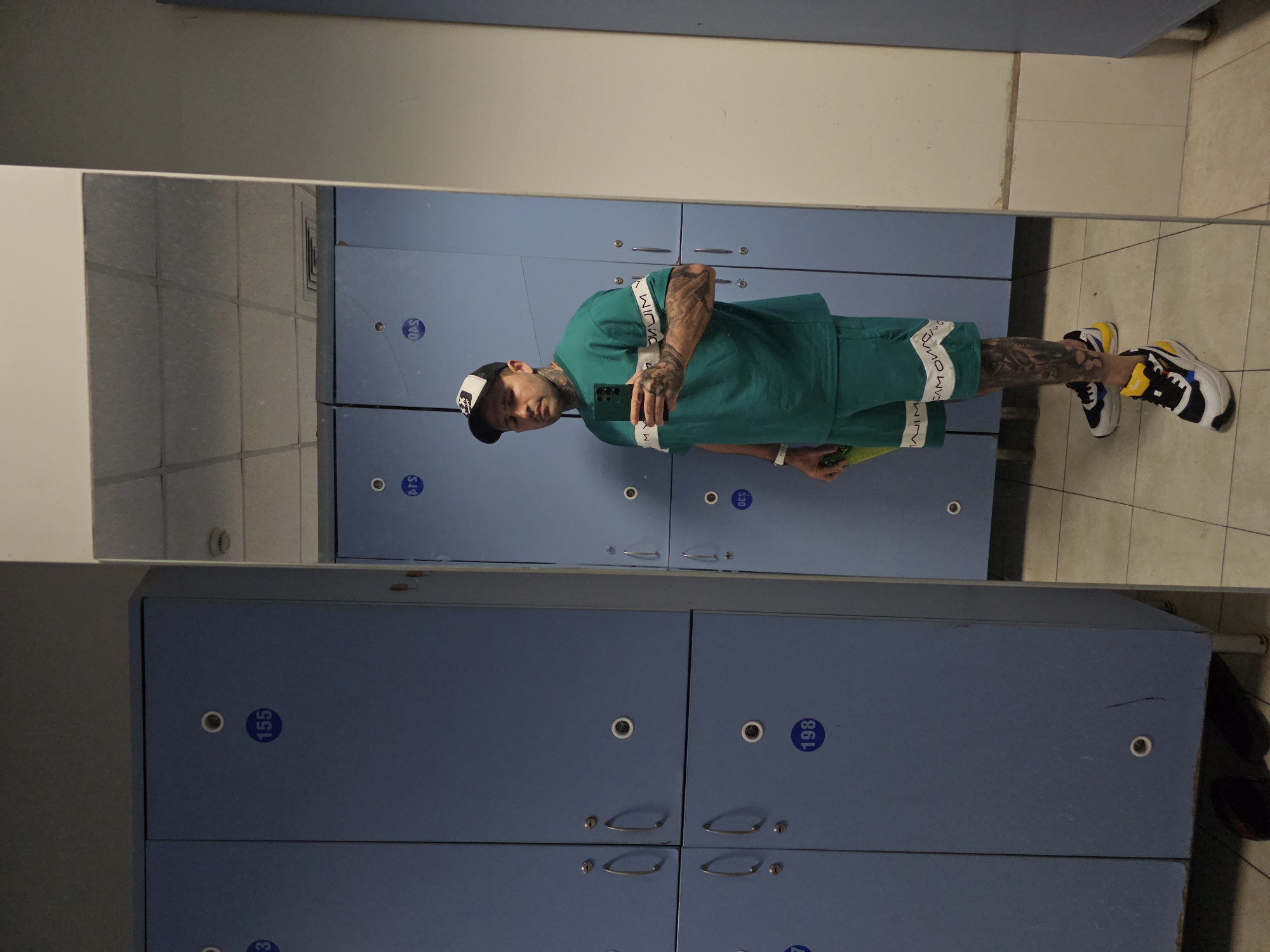The image depicts a man taking a mirror selfie in a locker room. He is wearing a green t-shirt with white stripes on the sleeves and matching shorts. He also has a black and white baseball cap on and is wearing colorful sneakers. The locker room is lined with blue lockers, some of which are labeled with numbers. The floor is tiled with square tiles. The man is holding a green bottle in his left hand and his phone in his right hand. The overall scene is consistent with a locker room in a gym or sports facility. The provided context that this image is from Stara Zagora, Bulgaria, does not offer any specific visual cues within the image itself to confirm this location.
