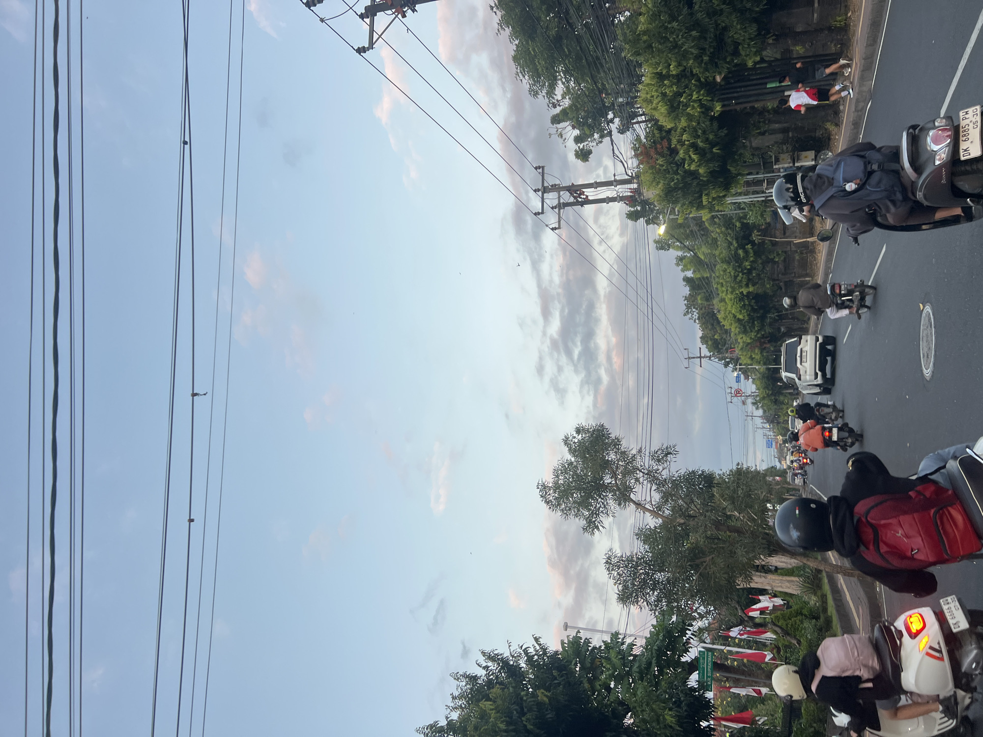 The image shows a street scene in Denpasar, Indonesia, likely during late afternoon or early evening, given the soft lighting and the pastel colors of the sky. Multiple motorcycles and a white SUV are traveling on a paved road. On the left side of the road, there are trees, some Indonesian flags, and a street sign indicating "W.R. SUPRATMAN". On the right side, more trees and electrical poles with numerous wires are visible. Several people are riding motorcycles, most wearing helmets. Two individuals are running on the sidewalk on the right side of the road. The sky is a blend of pale blue and light pink/gray clouds.