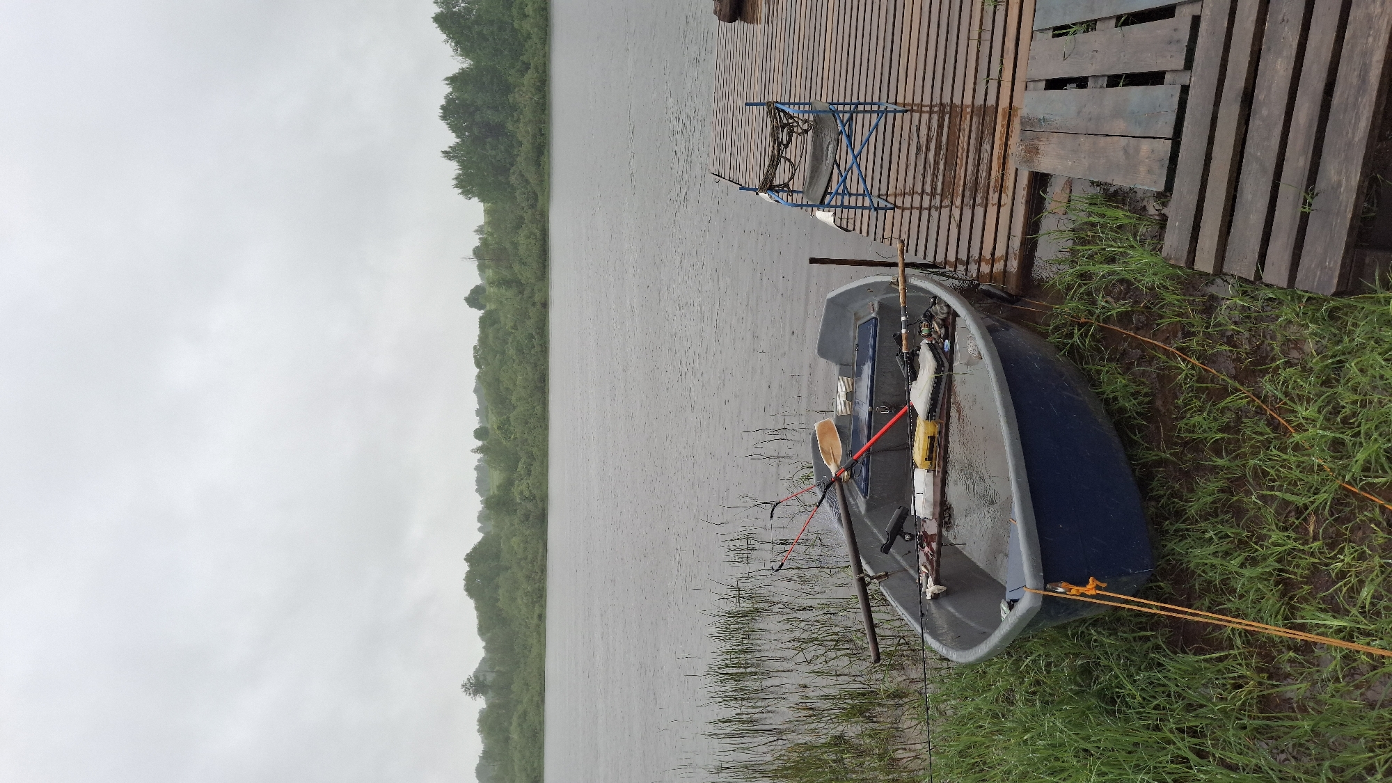 The image depicts a waterside scene in Sitno, Russia, under an overcast sky indicating rainy weather. A wide body of water, likely a river or lake, fills the mid-ground, its surface showing numerous ripples from falling rain. The distant bank is lined with a dense, dark green forest of deciduous trees. The near bank consists of muddy ground covered with vibrant green grass and sparse reeds.
In the foreground and mid-ground, a small, dark blue and gray open-top boat, approximately 3 meters in length, is partially beached on the muddy bank and moored alongside a wooden pier. The boat contains standing rainwater. Within the boat are multiple fishing rods, one with a red handle and an orange-bladed paddle across the gunwales. Inside, a small black fish finder is mounted, and various fishing supplies including several tackle boxes (yellow, white, clear) and other equipment are visible. An orange rope extends from the boat's bow into the left foreground, securing it to the bank.
To the right of the boat, a rustic wooden pier, constructed from horizontal, weathered planks, extends into the frame. The planks are wet, suggesting recent precipitation. A blue metal-framed folding chair with a dark fabric seat is positioned on the pier, draped with a coiled rope or fishing net. Further along the pier, a dark, cylindrical object rests on the planks. No individuals are present in the scene.