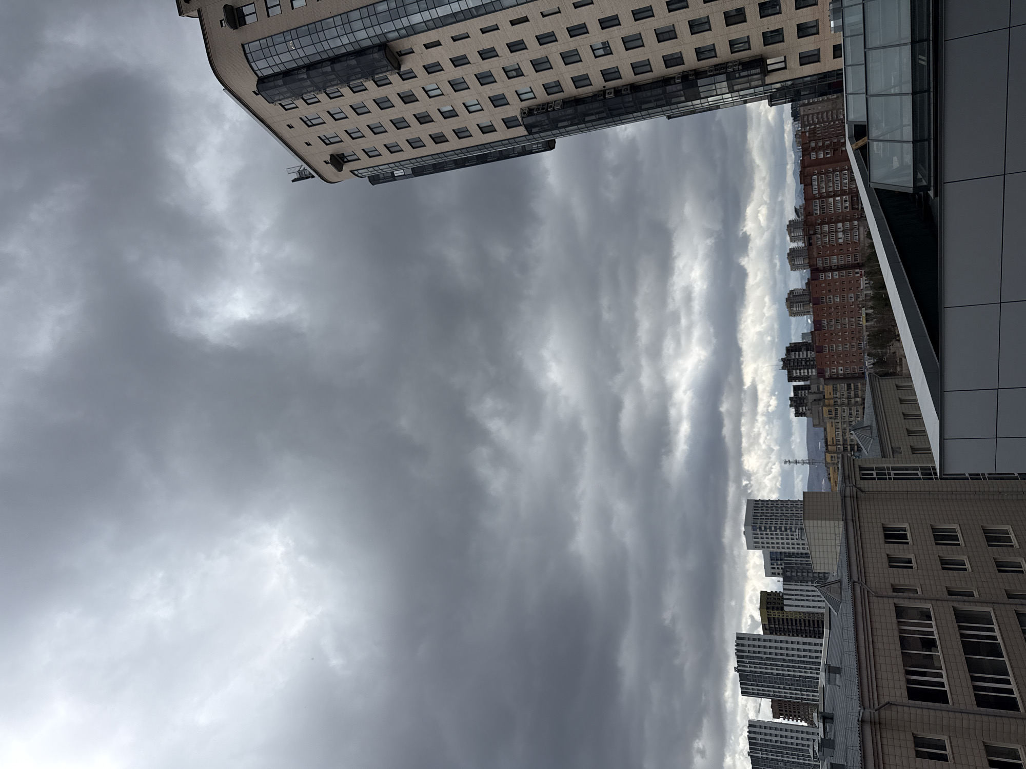 The image captures an urban scene under an overcast sky, likely in the city of Krasnoyarsk, Russia. The foreground is dominated by the grey, textured clouds. In the upper left and right corners, portions of modern high-rise buildings are visible. The architecture includes large windows and contemporary design. A few of the buildings in the distance appear to be older. There are no people visible. The composition of the image emphasizes the height of the buildings in relation to the sky. A tall communication tower is visible in the distance.