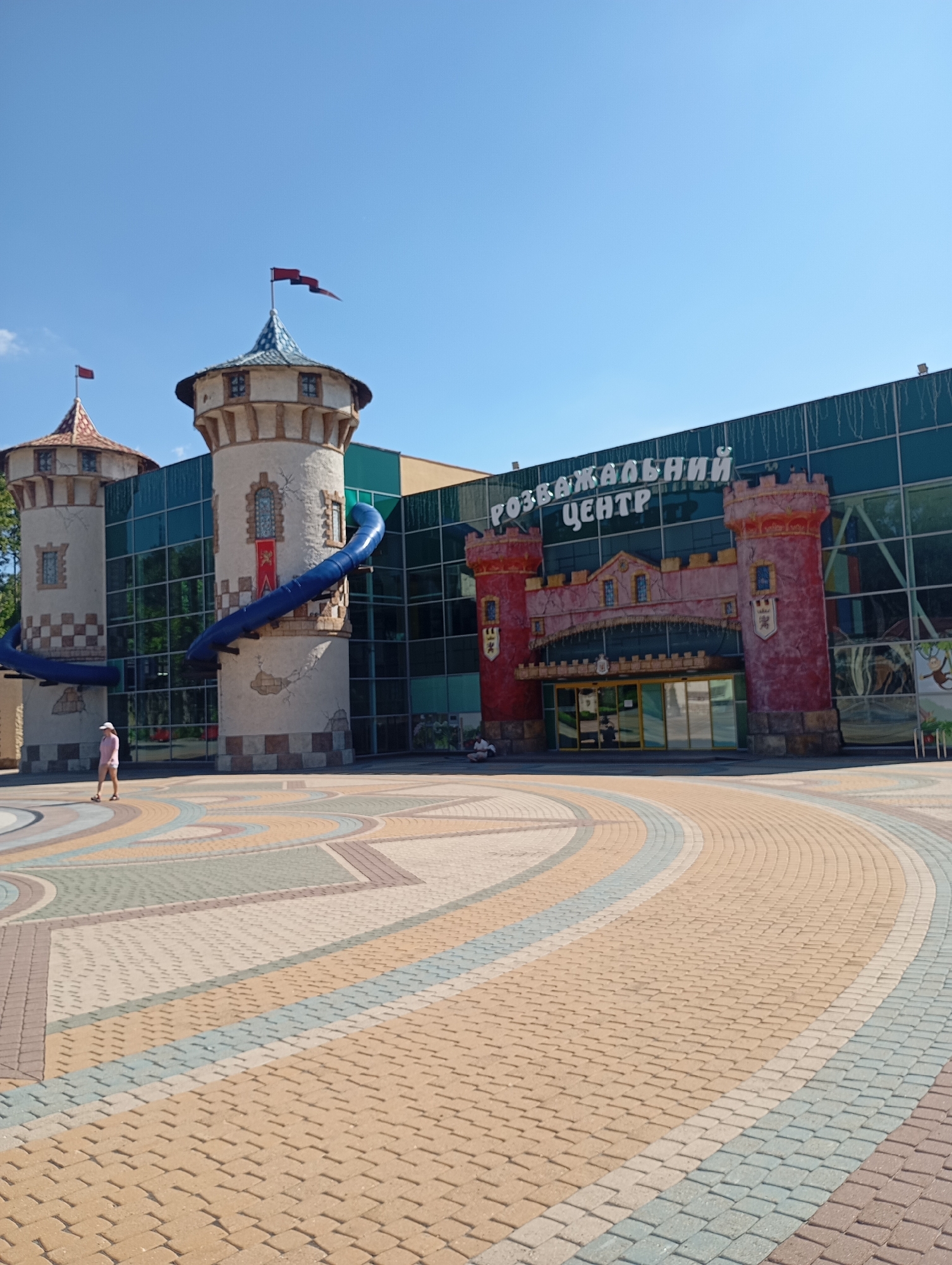 The image depicts the "Children's Development Center" (Дитячий Розвивальний Центр) in Kharkiv, Ukraine. The building's architecture is designed to resemble a medieval castle, with two prominent towers on the left and a larger, more elaborate castle facade on the right, featuring red and pink coloring and crenellations. A large blue slide is attached to one of the towers. The building has large glass windows, reflecting the clear blue sky and surrounding greenery. In front of the building is a wide, paved plaza with a decorative, colorful pattern of interlocking bricks. A lone person, wearing a pink shirt and shorts, walks across the plaza away from the camera, on the left side of the frame. The day is sunny, with a clear blue sky overhead.