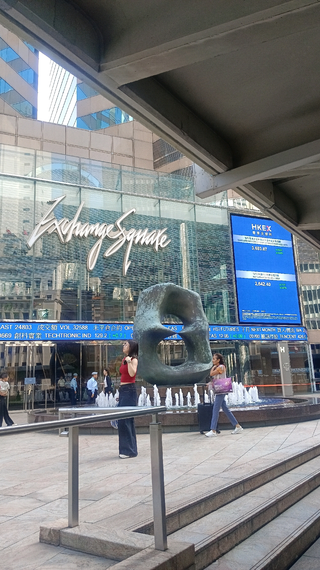 This image depicts the Exchange Square in Hong Kong, China, a prominent financial hub. The architectural style features a modern glass facade of a building displaying the "Exchange Square" logo in a flowing script. A large blue digital screen on the right side of the building shows stock market information from the Hong Kong Exchanges and Clearing Limited (HKEX). In the foreground, a large, abstract bronze sculpture with an opening in the center stands near a water fountain. Two women are visible in the mid-ground: one in a red top and dark jeans standing near the sculpture, and another in a patterned top and blue jeans walking with a suitcase and a purple shopping bag. Several other individuals, including security personnel and pedestrians, are present in the background near the building entrance. The scene is set on a paved plaza with stairs leading up from the foreground. The overall environment suggests a busy urban setting with a focus on finance and public art.