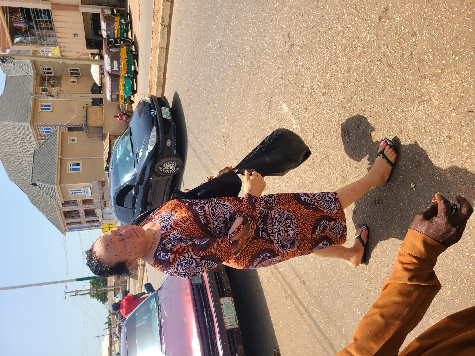 A woman walks along a paved road in Bukuru, Nigeria, under a clear, sunny sky. She is wearing a patterned orange and brown dress and flip-flops. She carries a black bag in her right hand. To her left, a maroon car is parked, and to her right, a black Peugeot car is driving on the road. In the background, several three-wheeled vehicles, commonly known as Keke Napep, are parked on the side of the road. Buildings, some appearing to be residential and commercial, line the street, and utility poles and wires are visible against the sky. The road surface is asphalt with visible cracks and debris. The overall impression is of a typical urban street scene in Nigeria.