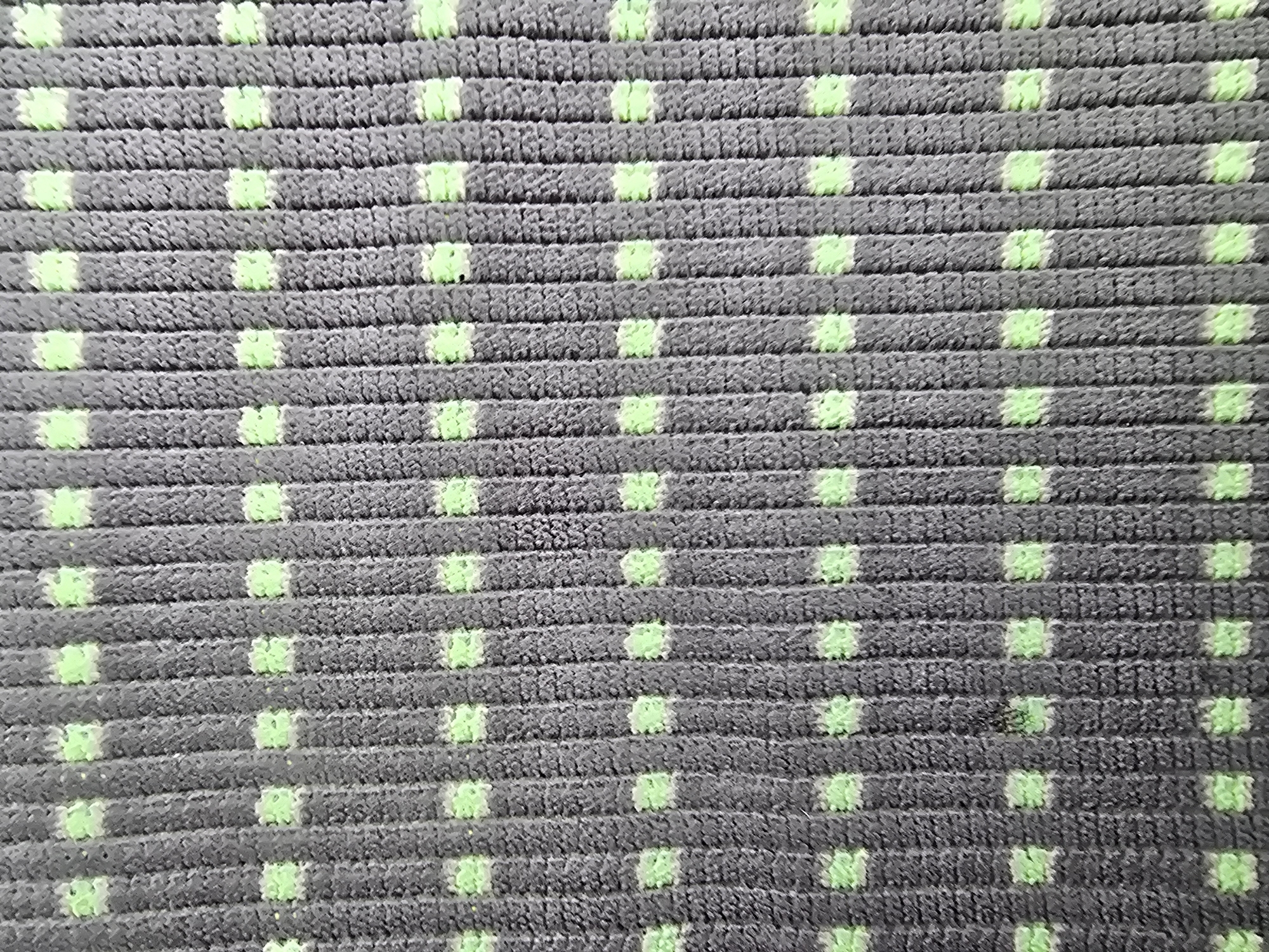 A close-up, overhead, full-frame shot shows a textured surface with a repeating pattern of vertical gray ribs and small, light green squares. The ribs are approximately one centimeter apart and have a fuzzy, corduroy-like texture. The green squares are evenly spaced, appearing in staggered rows and columns between the ribs. The lighting is neutral, with no distinct shadows, suggesting an indoor environment or diffused daylight. The image does not contain any identifiable landmarks or features that would allow for specific location attribution within Tbilisi, Georgia. No people, actions, or interactions are depicted.