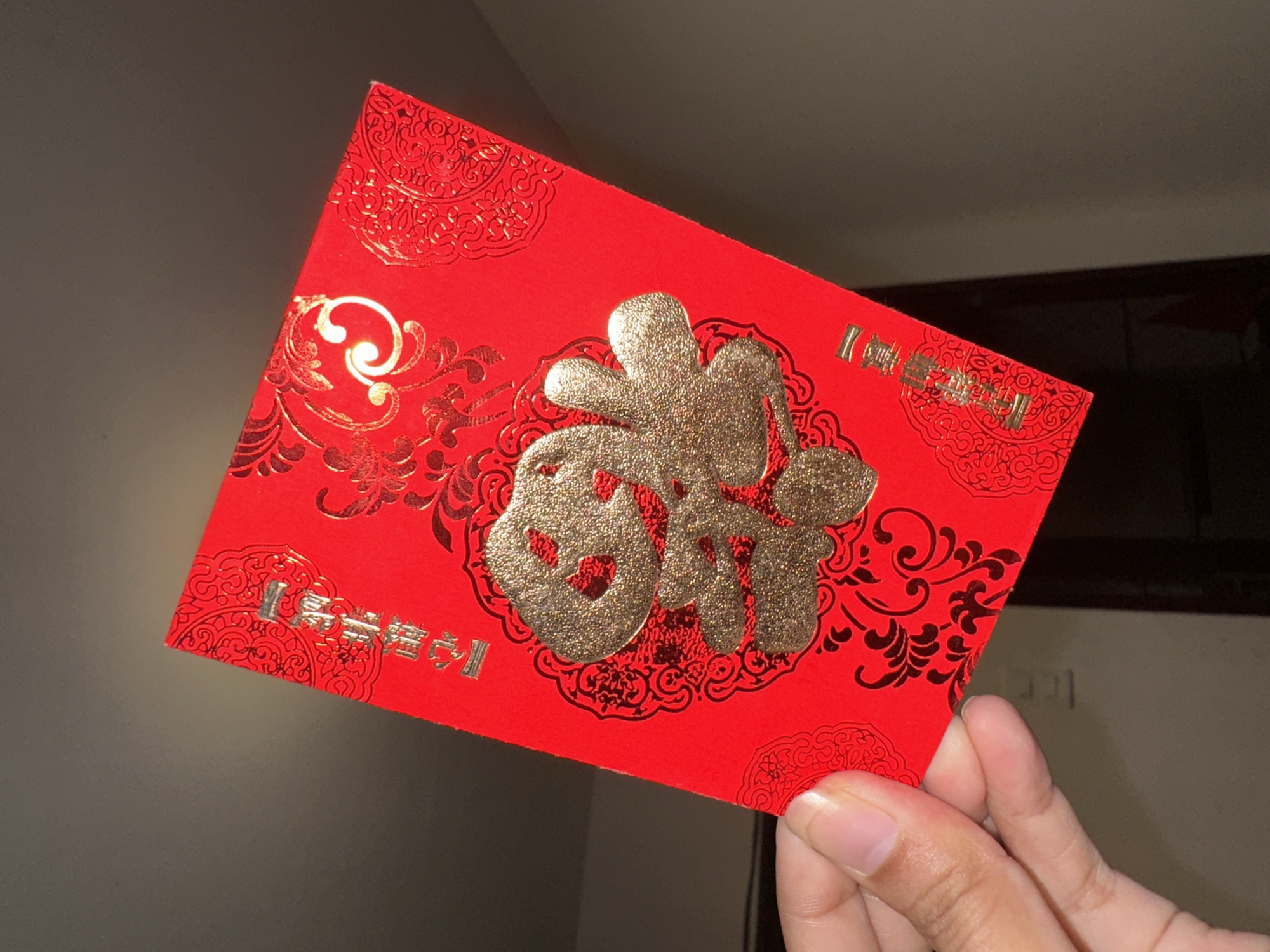 A close-up shot captures a human hand holding a vibrant red envelope, known as a hongbao or ang pao, in an indoor setting in Davao City, Philippines. The envelope is brightly colored red, adorned with intricate gold and black designs.
The most prominent feature of the envelope is a large, glittery gold Chinese character "福" (fú) centrally located. This character signifies 'fortune', 'blessing', or 'good luck'. Surrounding this central character are ornate black and gold scrollwork and floral patterns, enhancing the traditional and festive aesthetic of the envelope.
In the top left corner of the envelope, smaller gold characters read "恭喜發財" (gōng xǐ fā cái), a common greeting wishing 'prosperity and wealth'. A smaller, identical "福" character is also visible in the bottom right corner, partially obscured by the thumb of the hand holding the envelope.
The hand, with light skin tone and visible fingernails, gently grasps the envelope from its bottom right edge. The background is dark, indistinct, and out of focus, indicating an indoor environment where direct lighting highlights the shimmering gold details of the envelope. The image depicts a cultural practice of giving monetary gifts in red envelopes, symbolizing good wishes and blessings, often observed during celebrations like Chinese New Year, weddings, or birthdays within Chinese and Chinese-Filipino communities.