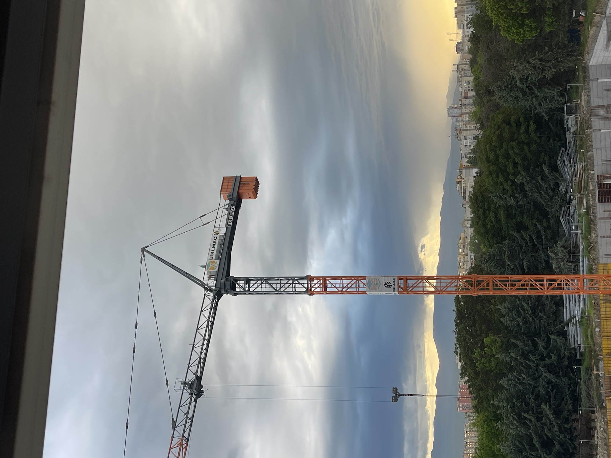 The image shows a construction crane with an orange and gray arm, branded "EDILMAC", visible from a building window. The crane is holding a small load of orange brick-like materials. In the background, there is a cityscape with buildings and trees. A mountain range is visible on the horizon. The sky is cloudy with varying shades of gray and a hint of yellowish light near the horizon. The location appears to be Plovdiv, Bulgaria. A construction site with steel beams is directly below the crane.